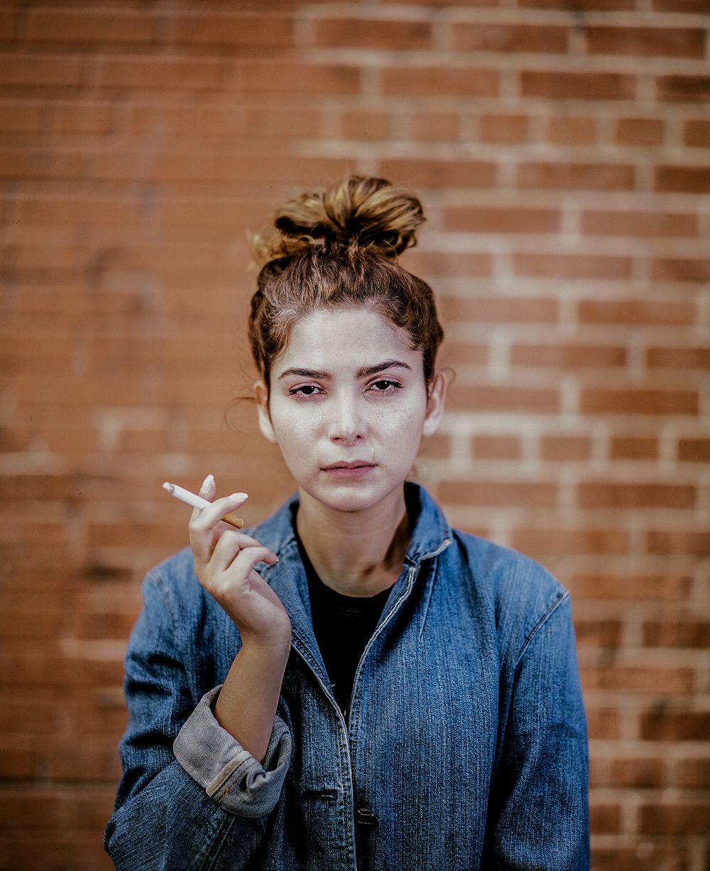 Person in denim jacket holding cigarette against brick wall background.