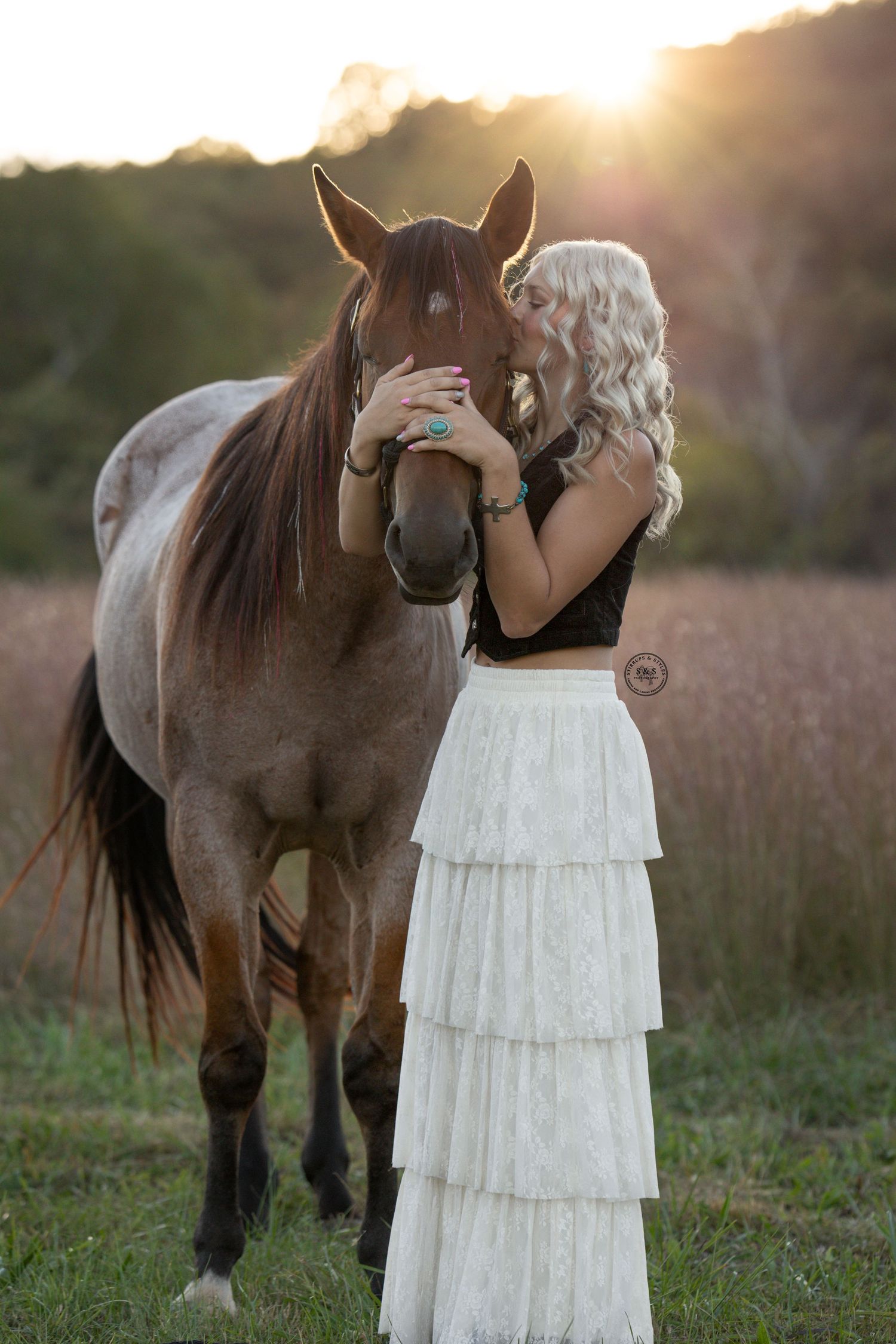 A woman in a white tiered dress shares a tender moment with a brown horse at sunset in a rustic field setting.