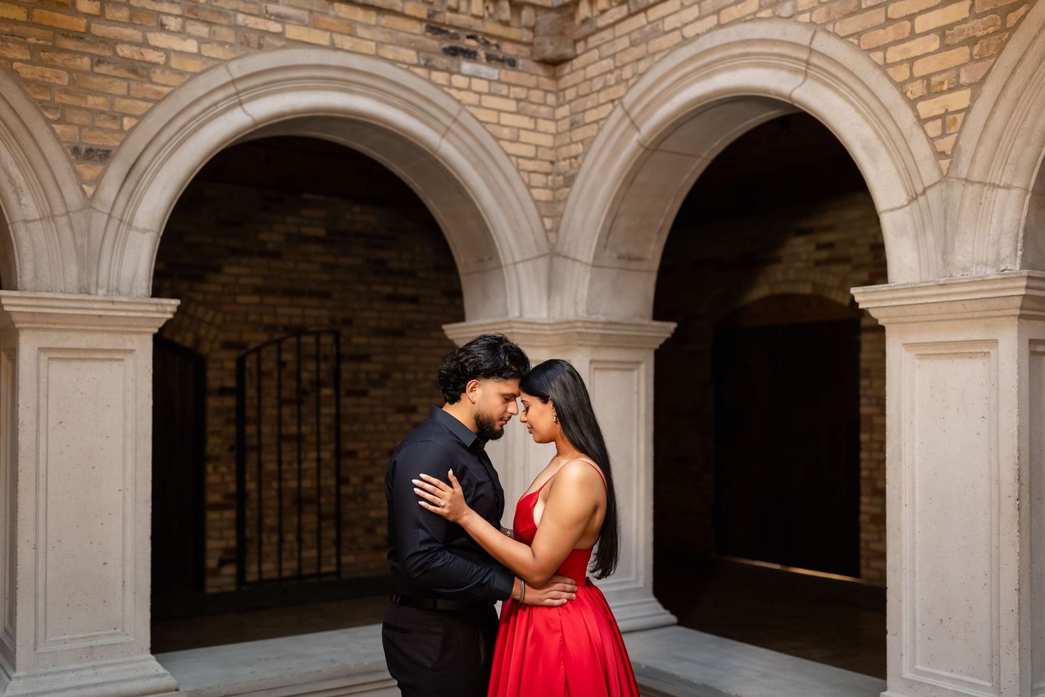 A couple in formal attire embraces under stone archways during an engagement photo session.