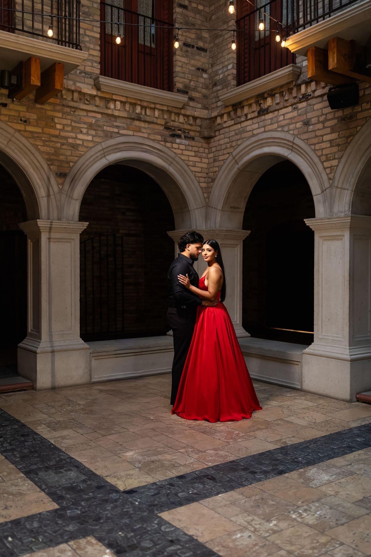 A couple in formal attire share an intimate moment under stone archways in an elegant courtyard setting.