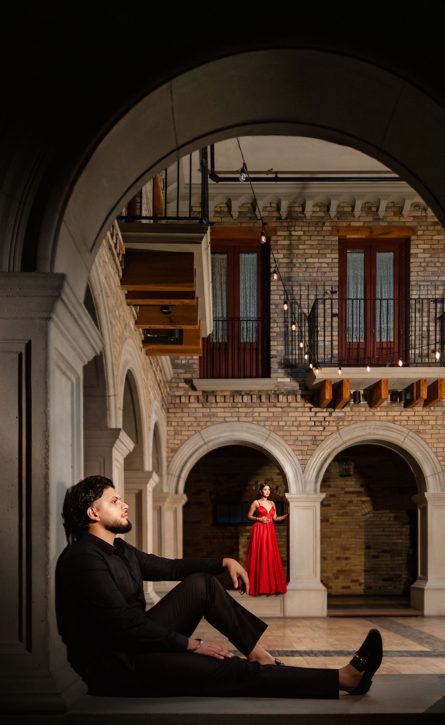 Dramatic indoor architectural scene with arched columns and brick walls featuring two people in contrasting black and red attire.