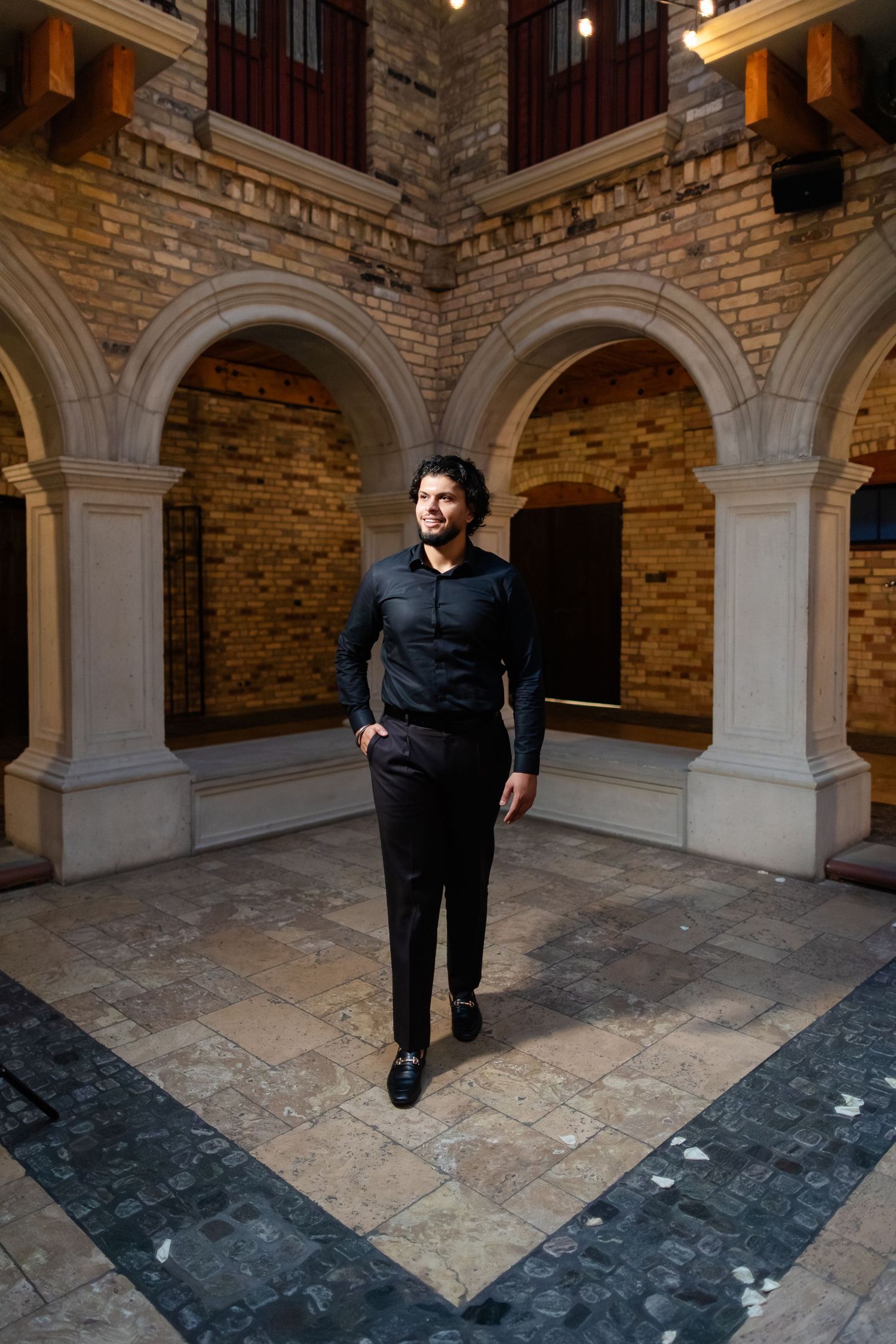 A person in formal black attire stands in an elegant stone courtyard with arched columns and brick walls.