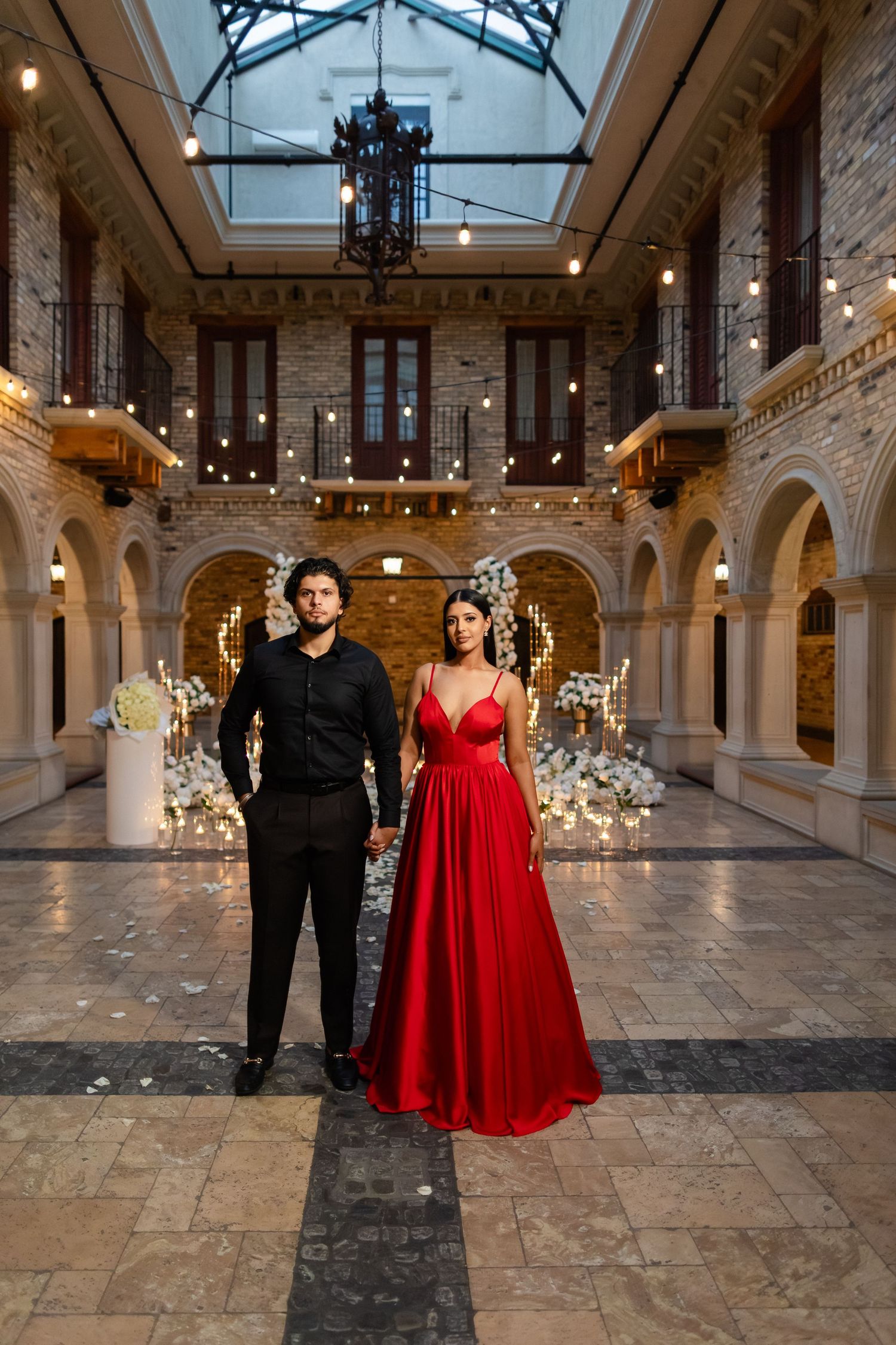 Elegant couple poses in formal attire inside historic stone building with arched architecture and twinkling lights.