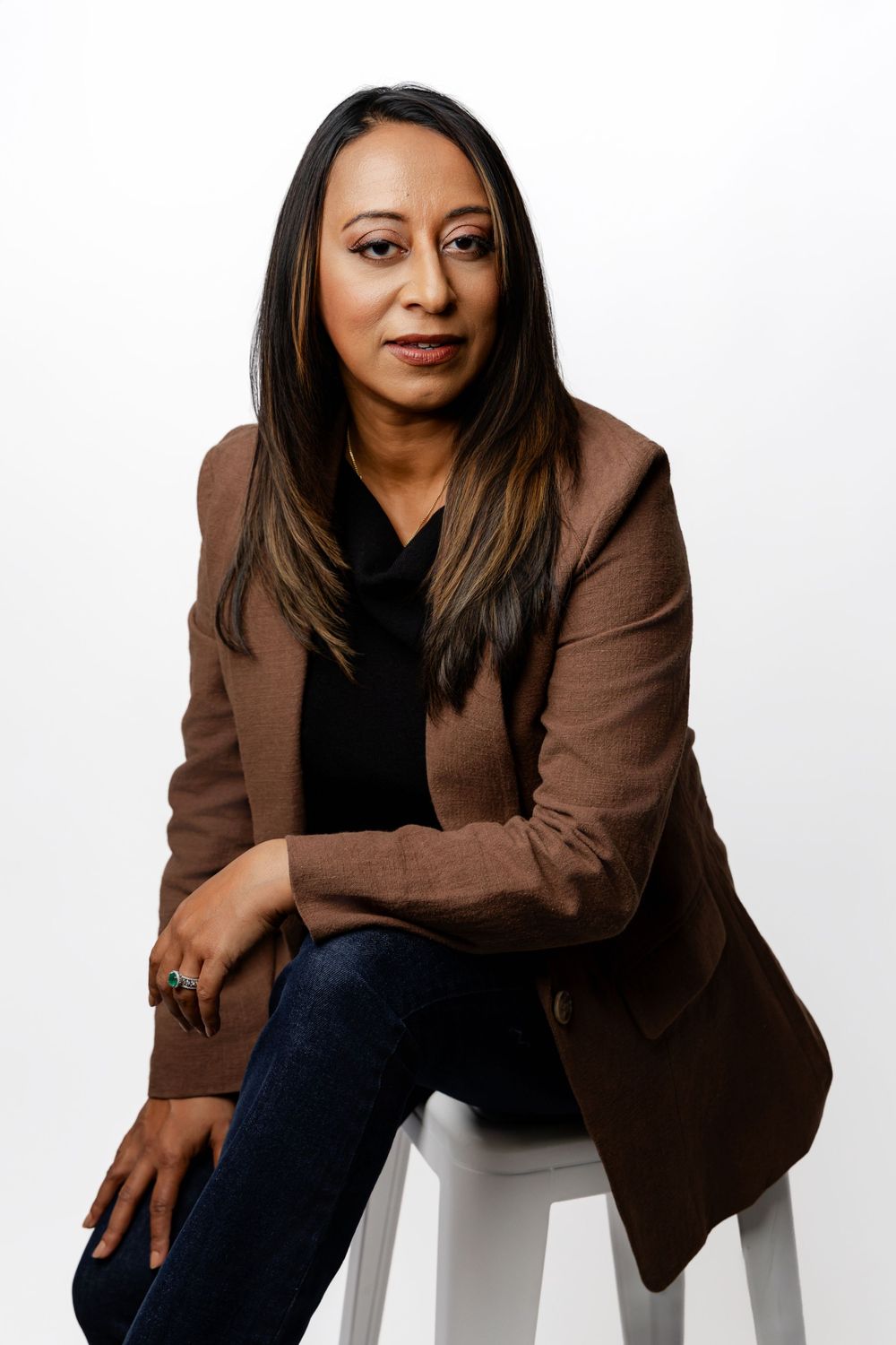 Professional portrait of someone in a brown blazer and black top seated on a white stool against white background.