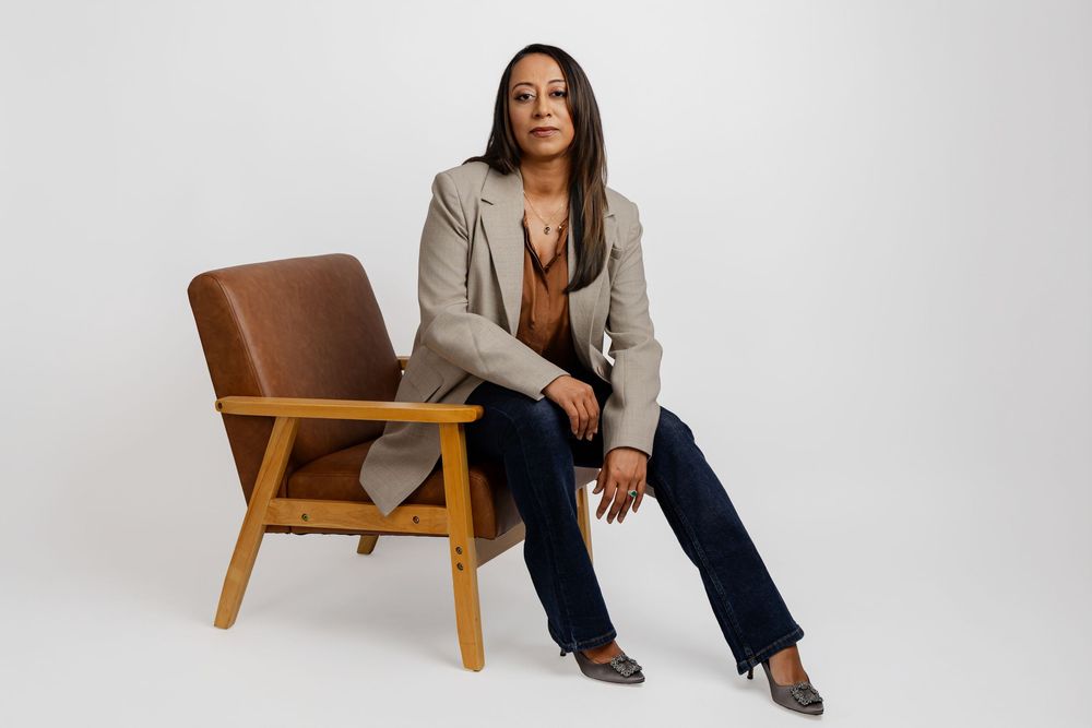A person in a beige blazer and dark pants sits casually on a wooden chair against a white background.