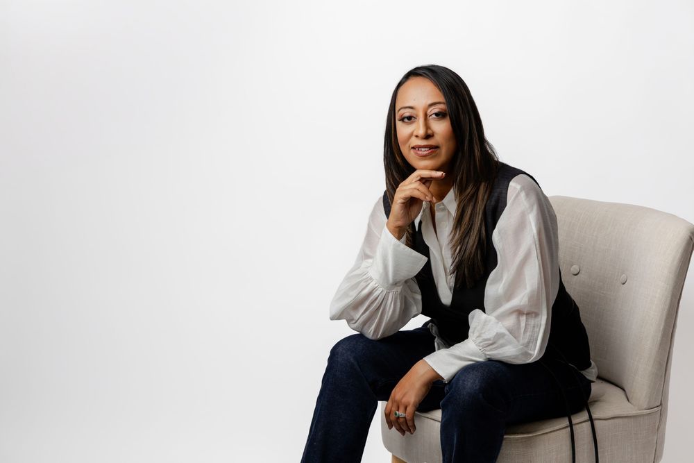 Professional woman in white blouse and dark pants sits elegantly on a white chair in studio setting.