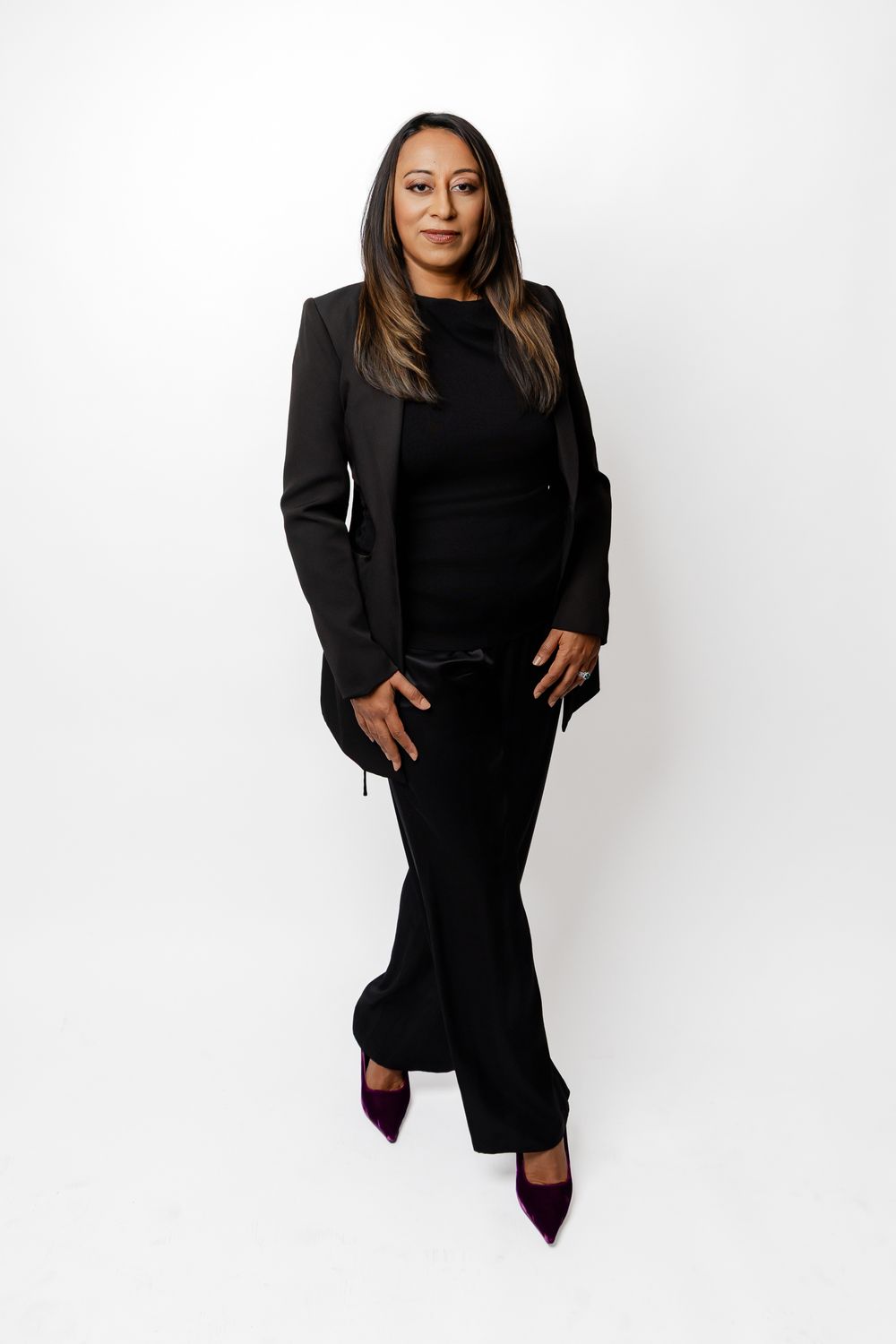 Professional studio portrait of someone wearing an all-black outfit with a blazer and burgundy heels against white background.