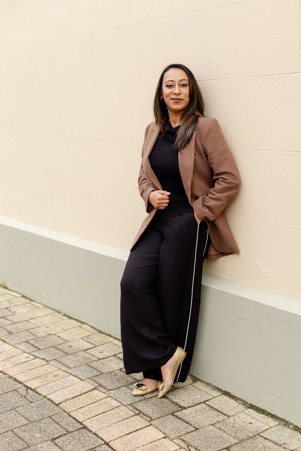 A fashionable outfit featuring a black dress paired with a brown blazer against a light colored wall.
