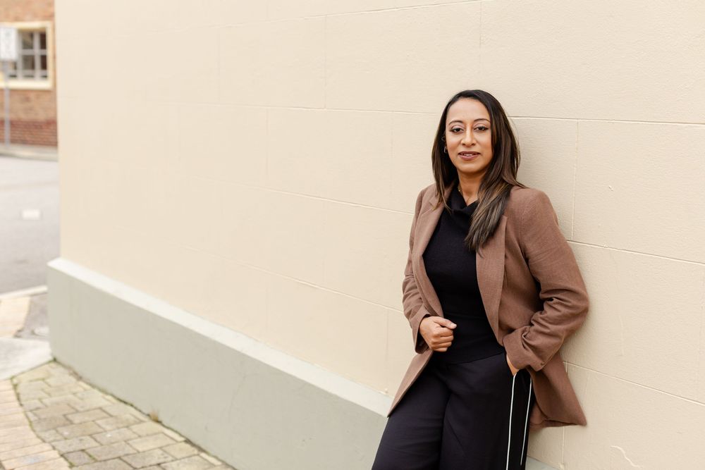 A person wearing a brown blazer and black outfit poses confidently against a white wall in an urban setting.