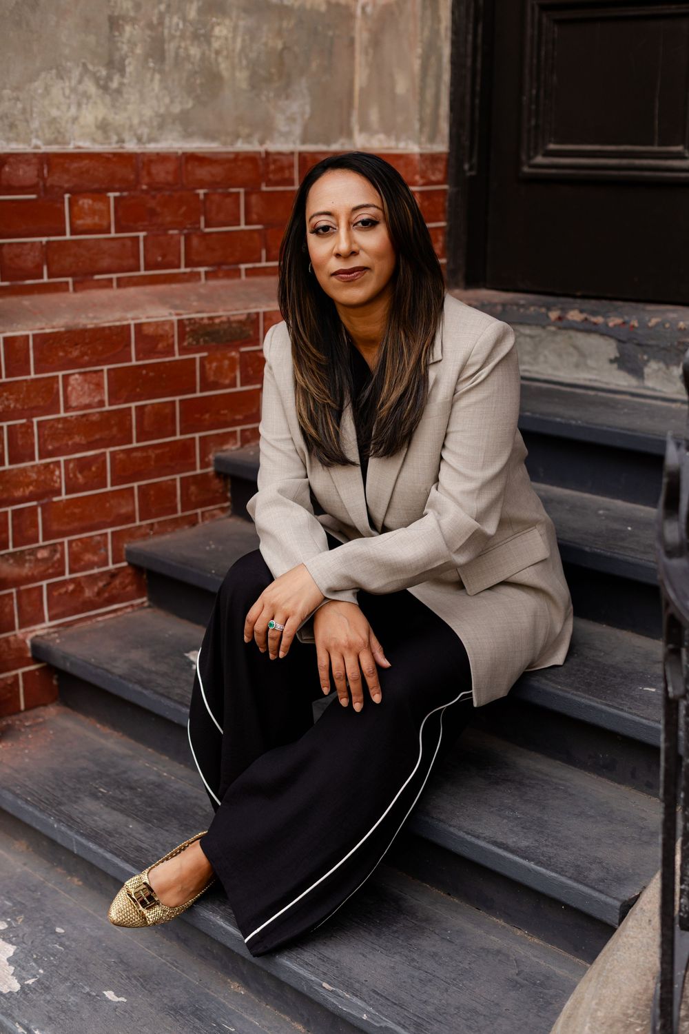 A person in a light blazer and dark pants sits casually on wooden steps against a brick wall background.