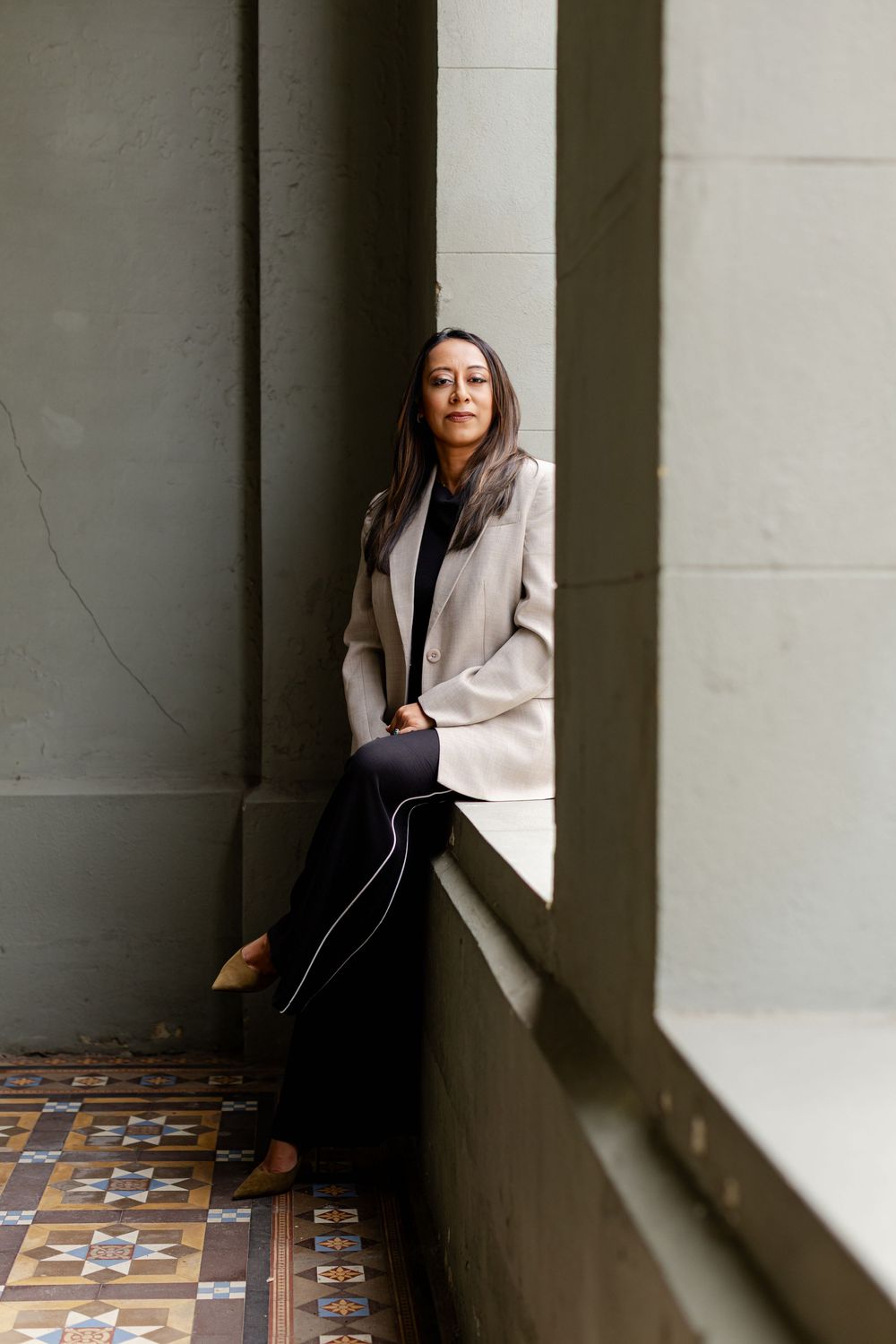 Person in beige blazer and black pants sits on window ledge with ornate tiled floor in architectural setting.