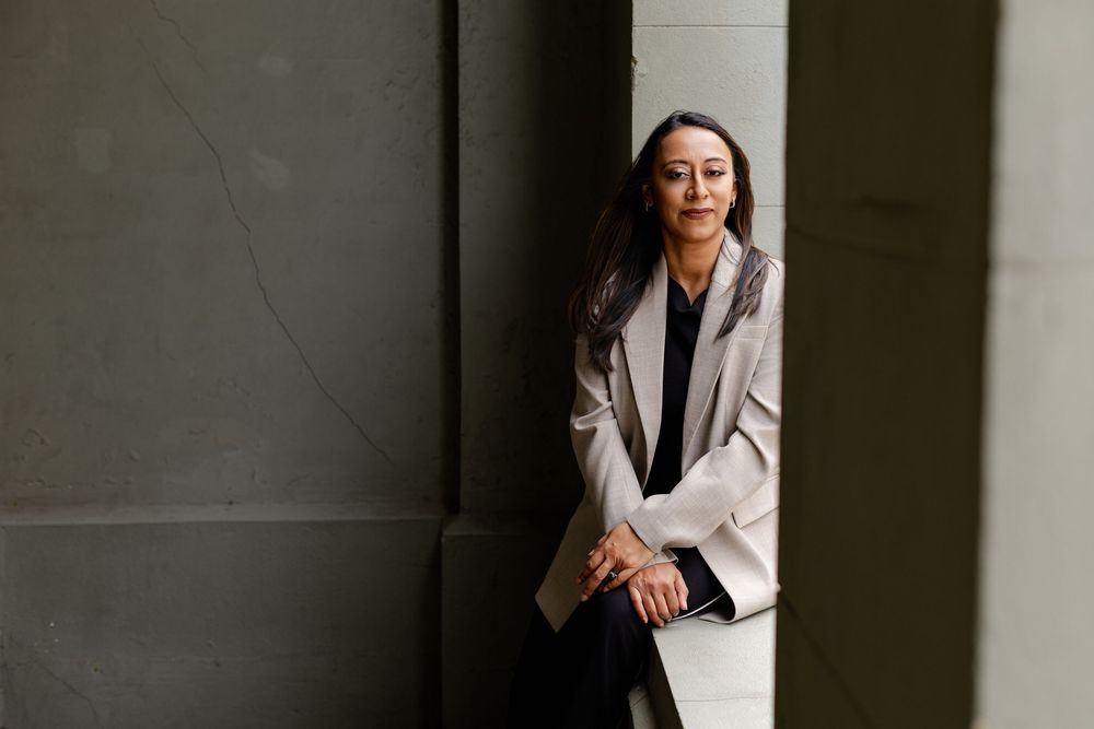 Professional wearing a white blazer poses in dramatic lighting against a dark wall with strong shadows.