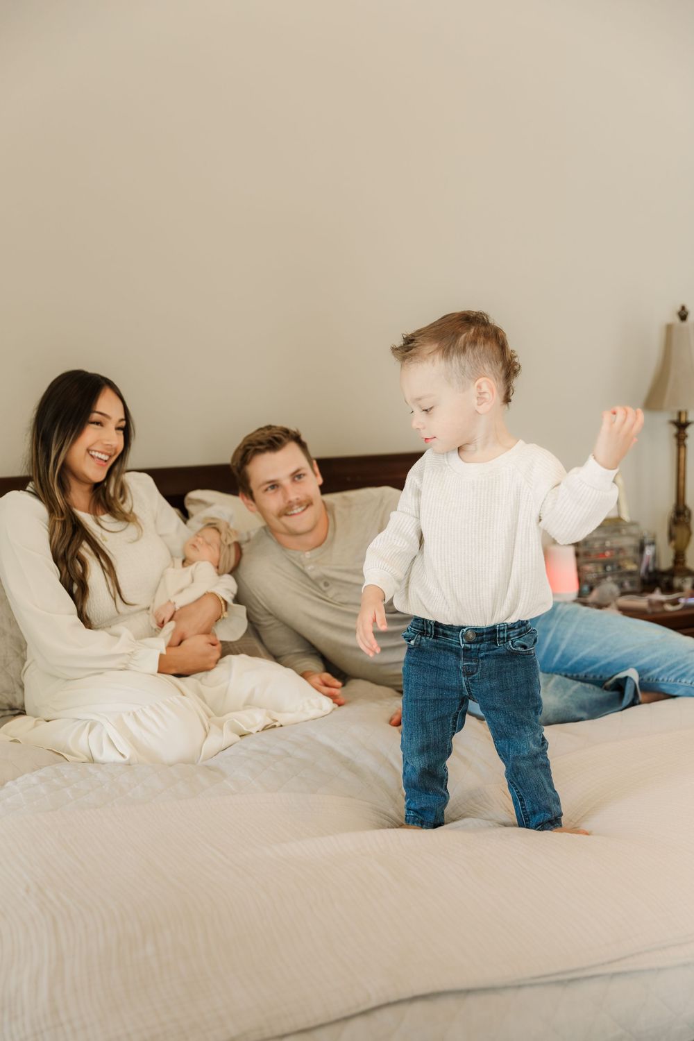 A family relaxes together on a white bed as a toddler in jeans stands and plays on the mattress.