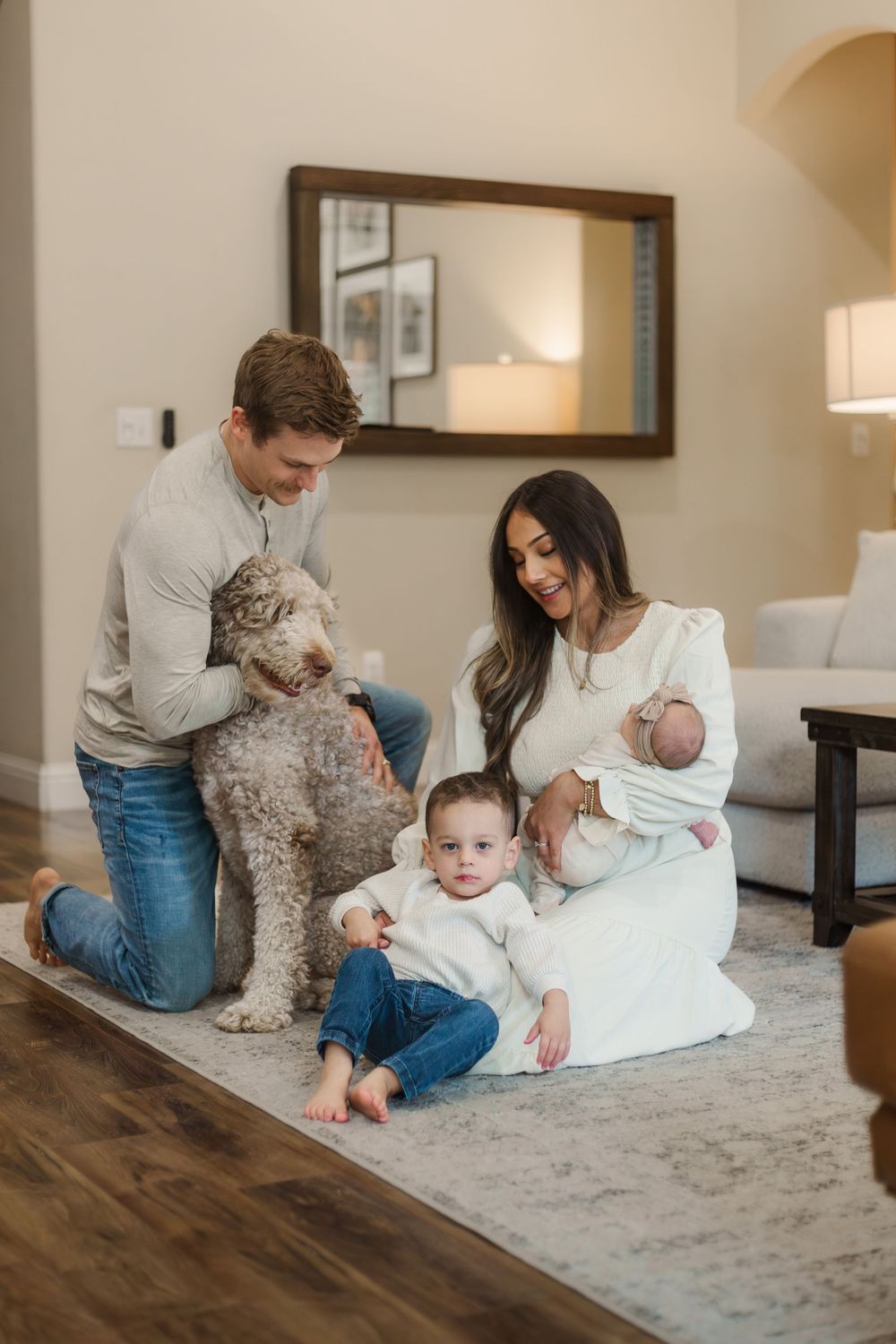 A family sits on the floor with their curly-haired dog and newborn baby in a cozy living room setting.