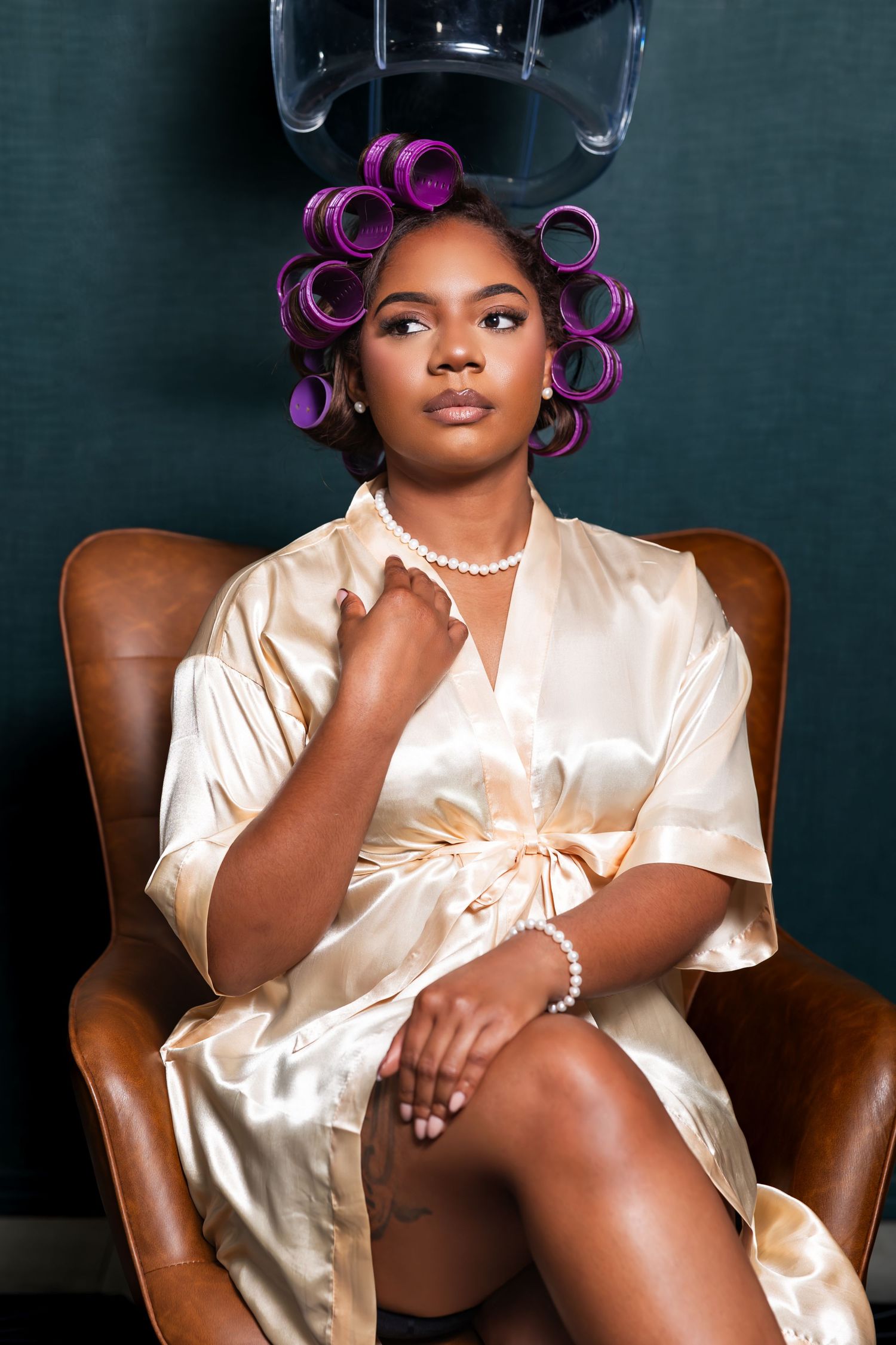 A figure in a cream silk robe and purple hair rollers poses elegantly on a brown leather chair in a studio setting.