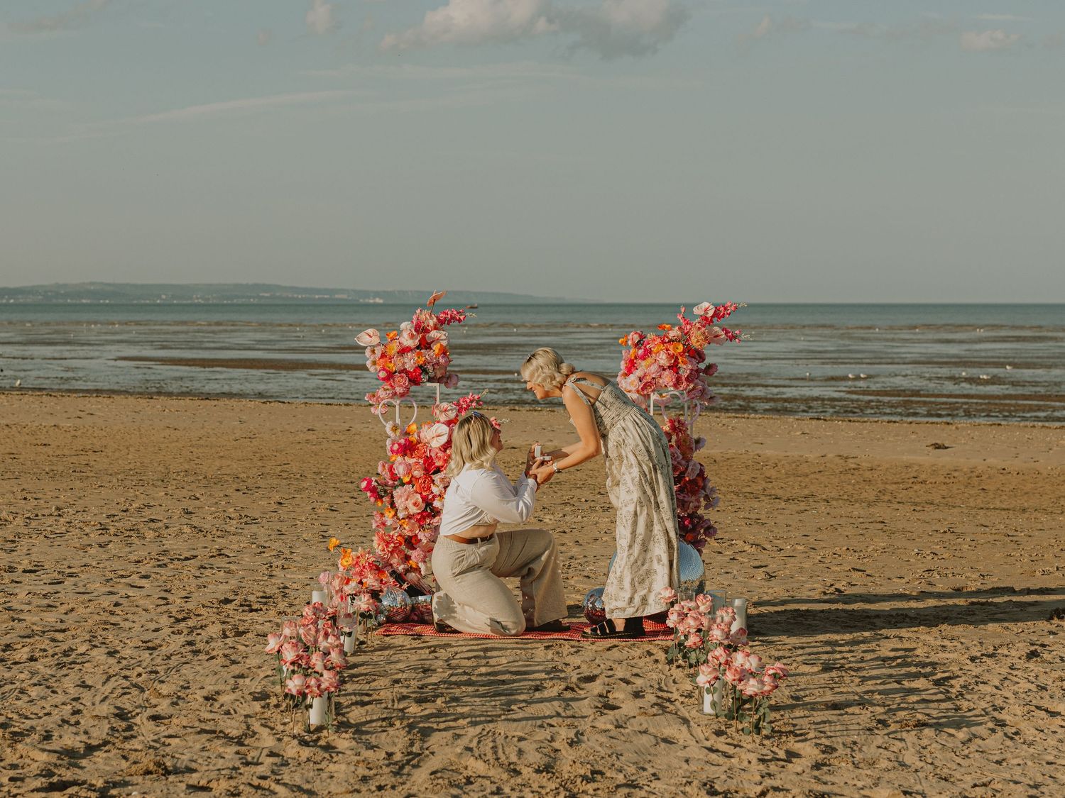 A romantic beach setup with coral floral arrangements and decor against a sandy shoreline at sunset.