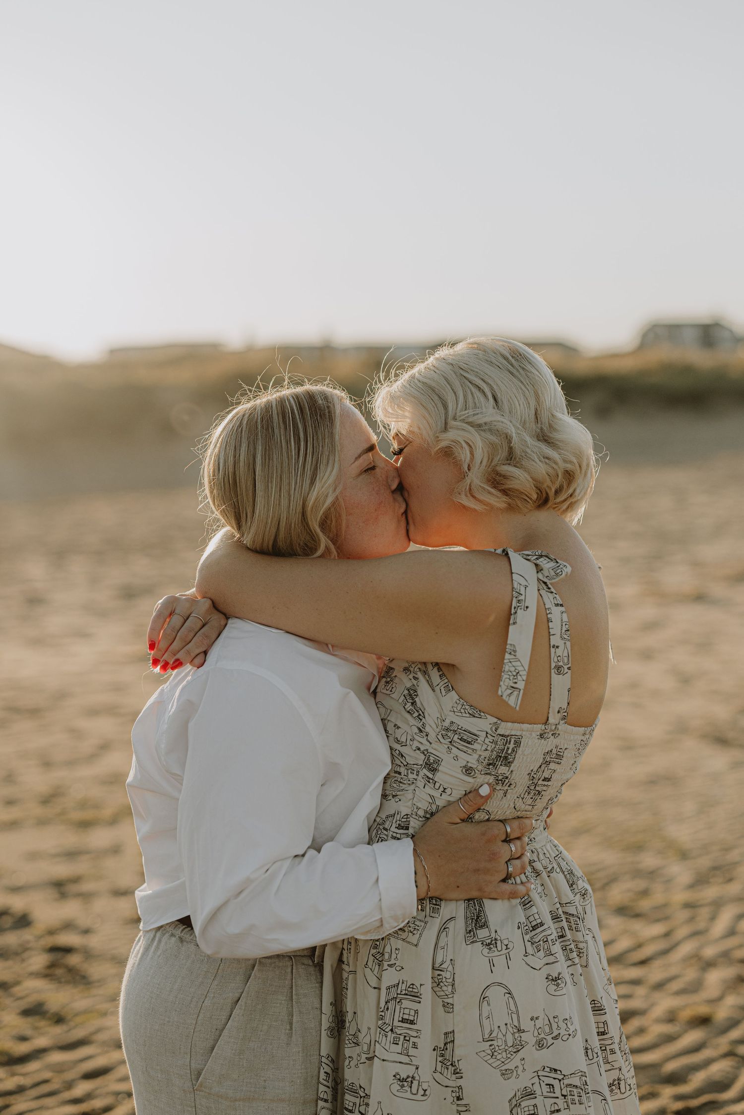 Two people in white and floral outfits embracing on a sandy beach.