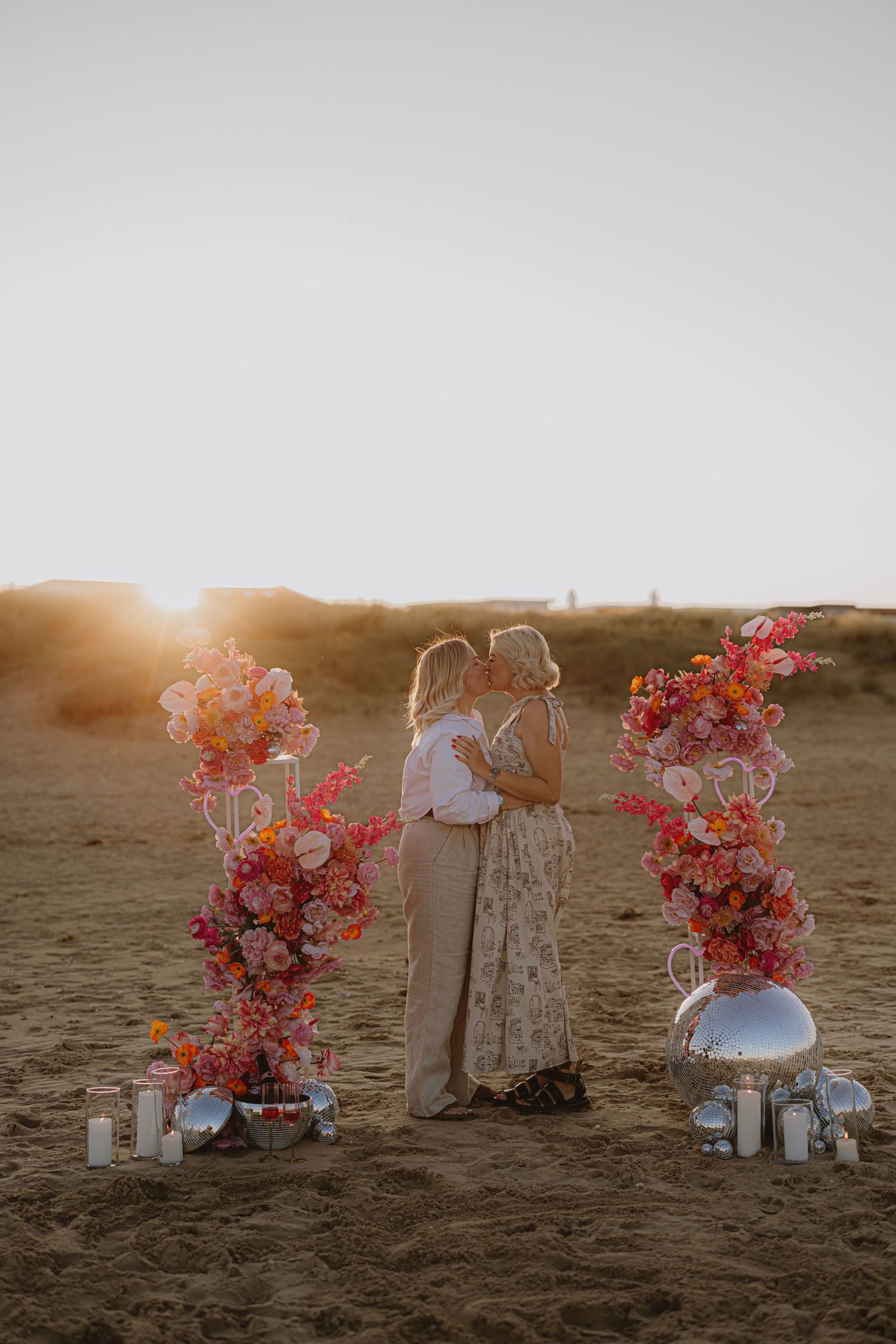 Beach ceremony decor with coral floral arrangements and white furniture at sunset.