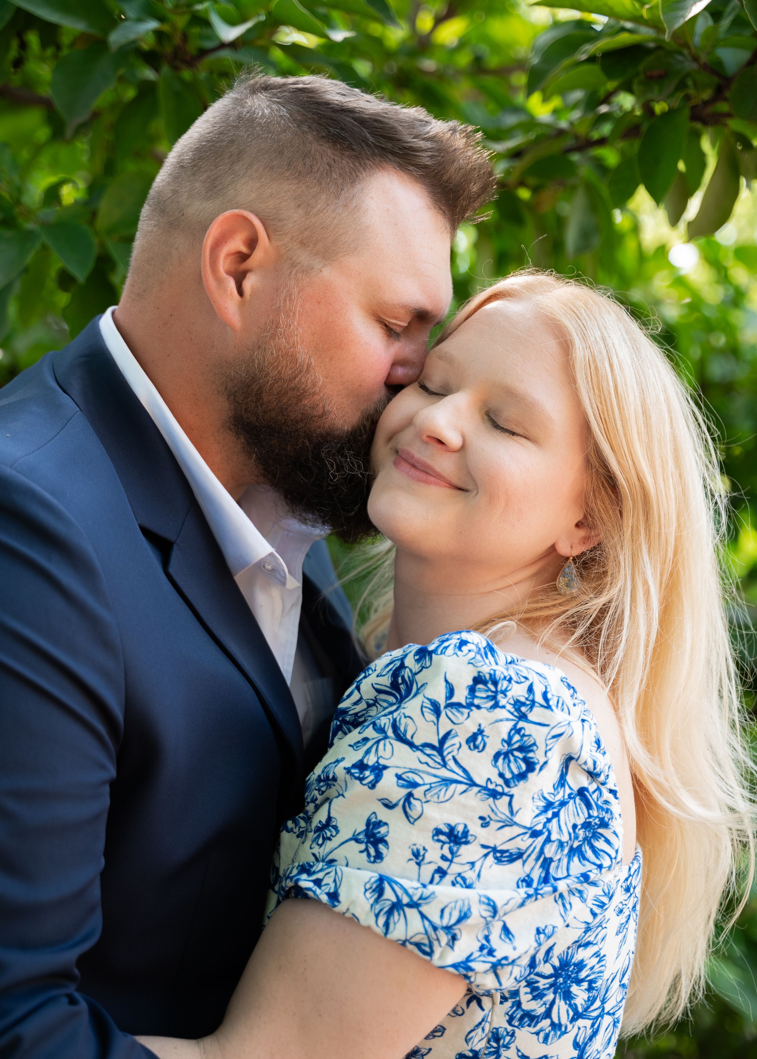 close up of man kissing womans cheek as she dreamily smiles with her eyes closed. Powell gardens greenery in the background