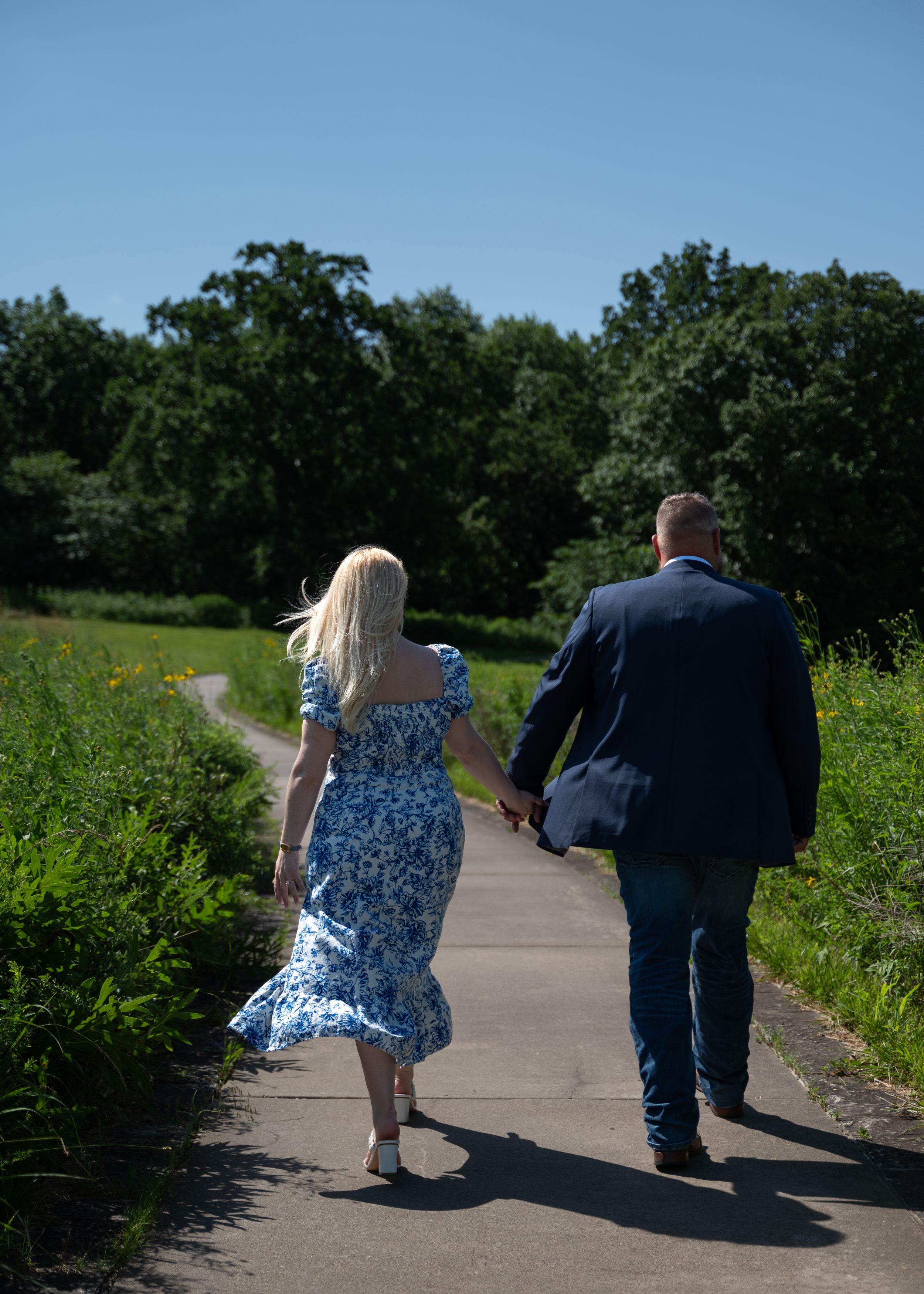 A couple walking hand in hand down a paved trail surrounded by green summer foliage.