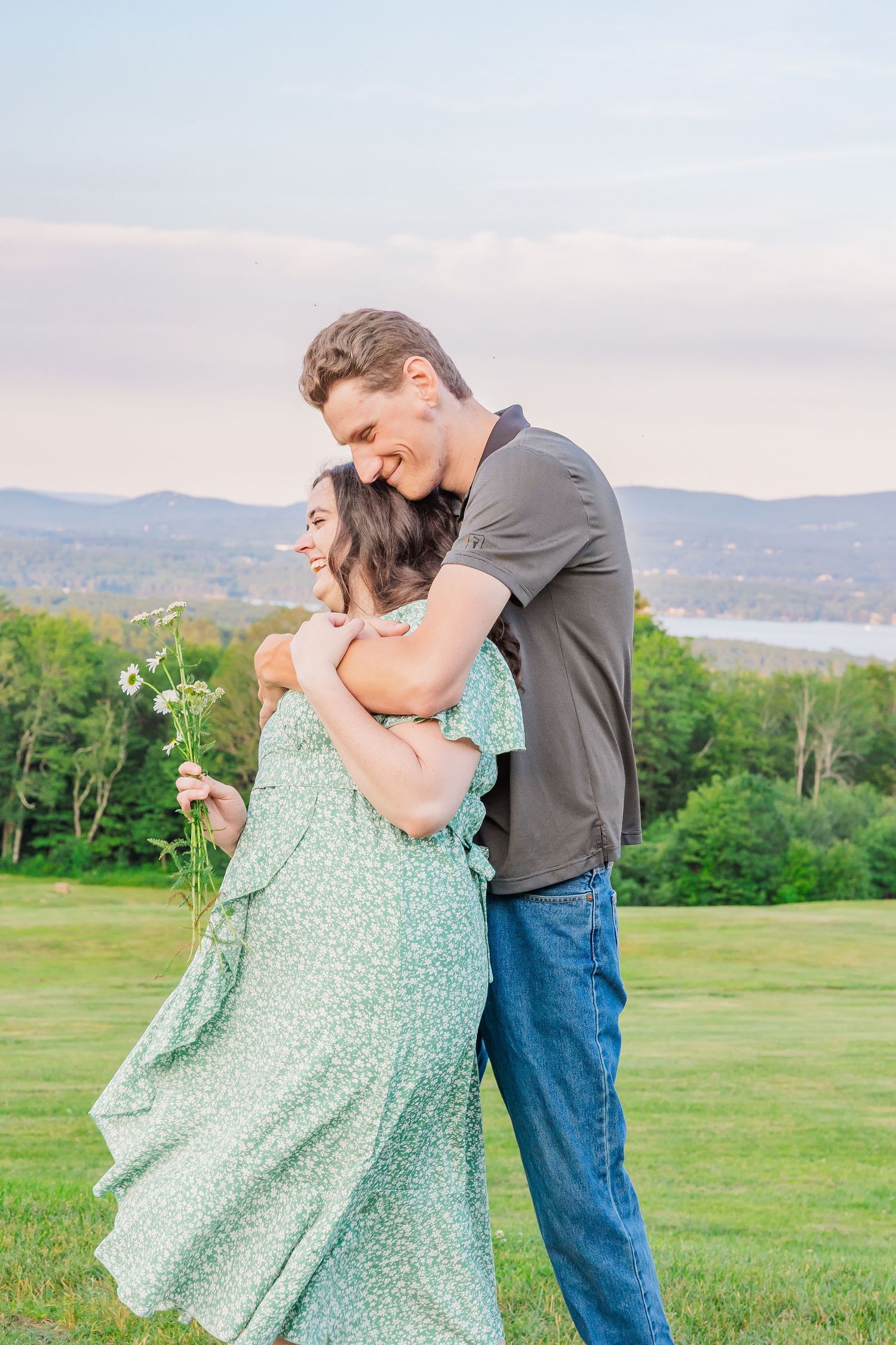 A couple in a green floral dress and dark shirt embrace lovingly on a hilltop meadow with mountain views at sunset.