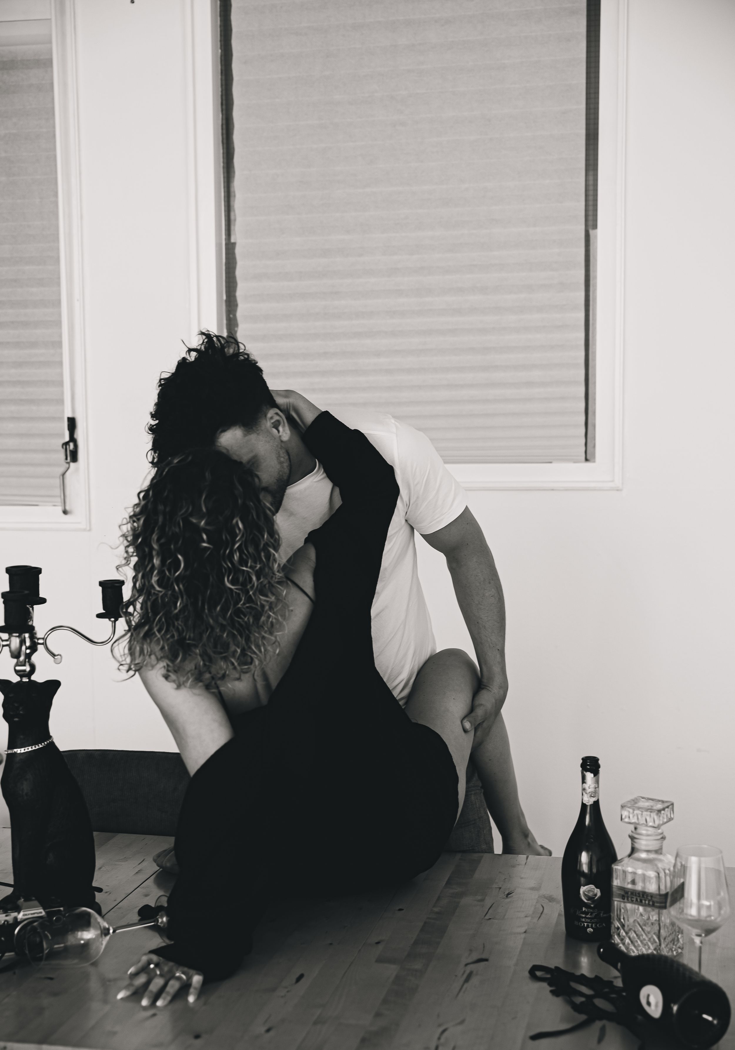 Black and white artistic photograph showing silhouette seated on floor near window with wine bottle and glasses.