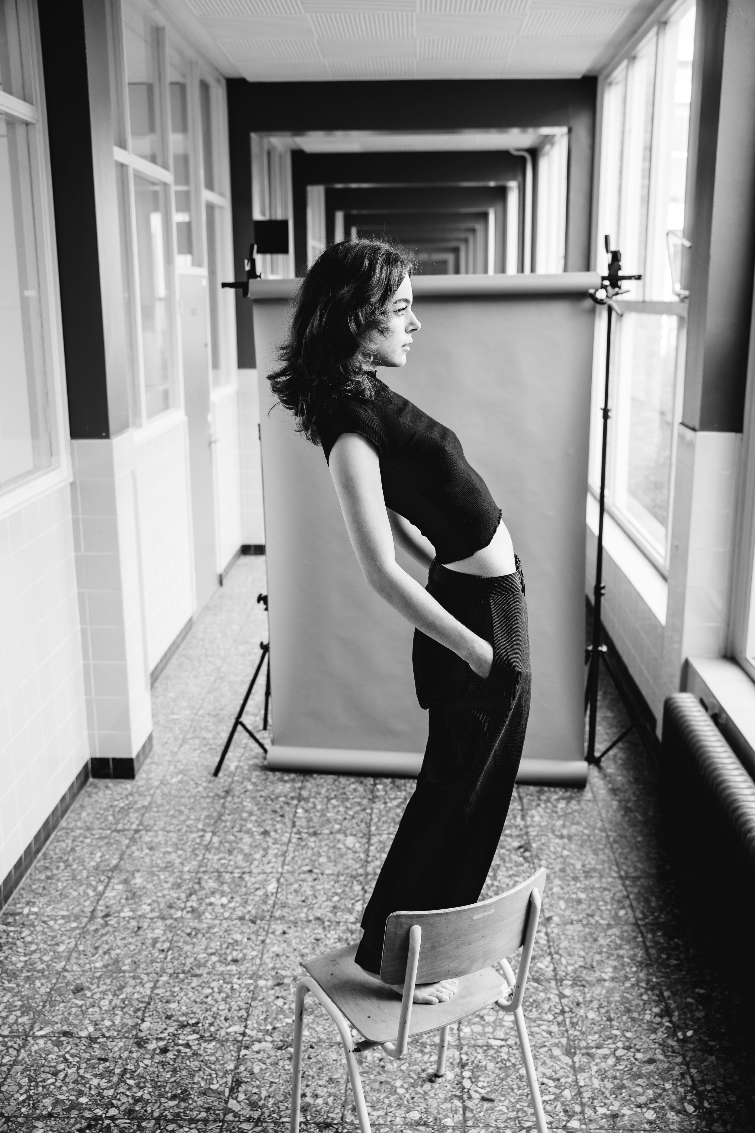 Black and white portrait photograph of a figure standing on a chair in a long hallway with dramatic lighting.