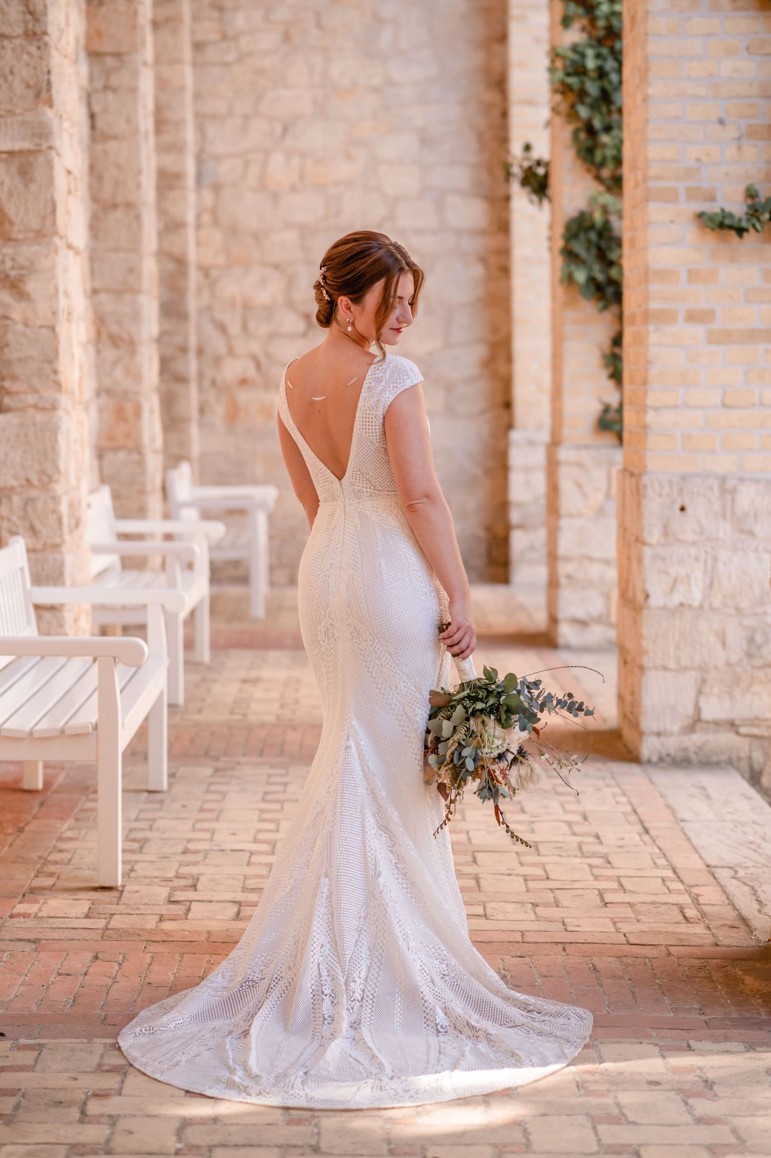 Elegant white lace wedding dress with open back design and trailing train on stone patio with ivy-covered columns.