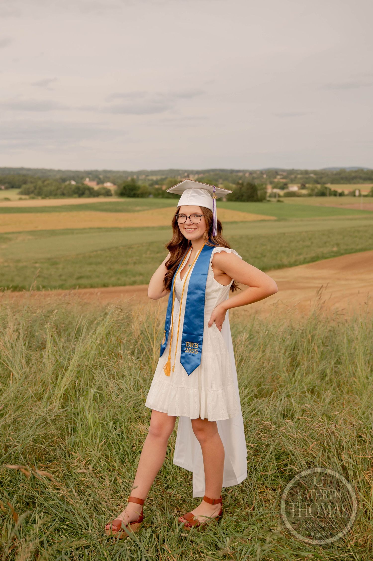 Graduate in white dress and blue sash poses in grassy field with rolling hills in background on sunny day.