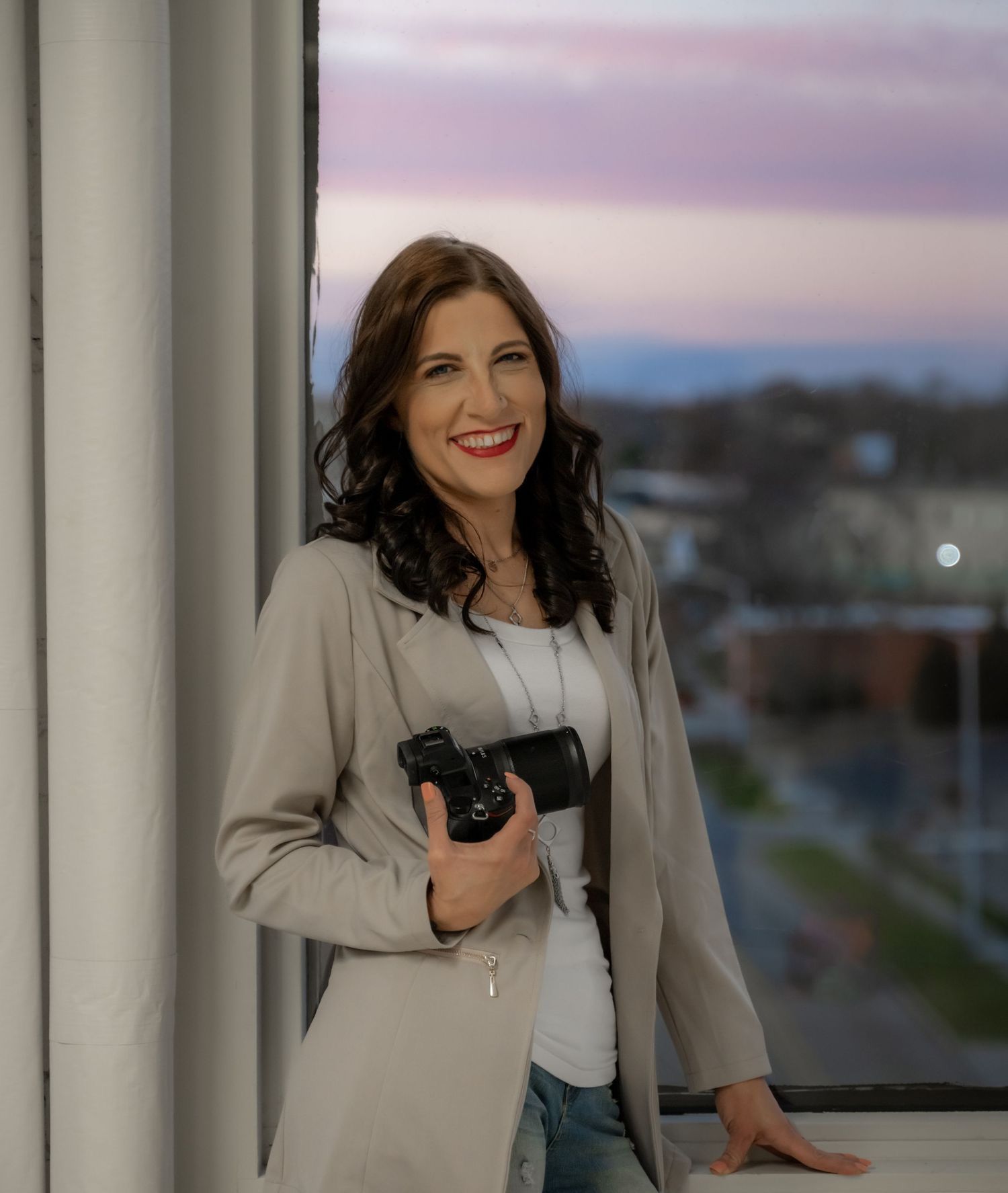 Professional photographer holding camera while leaning against pillar at dusk with cityscape in background.