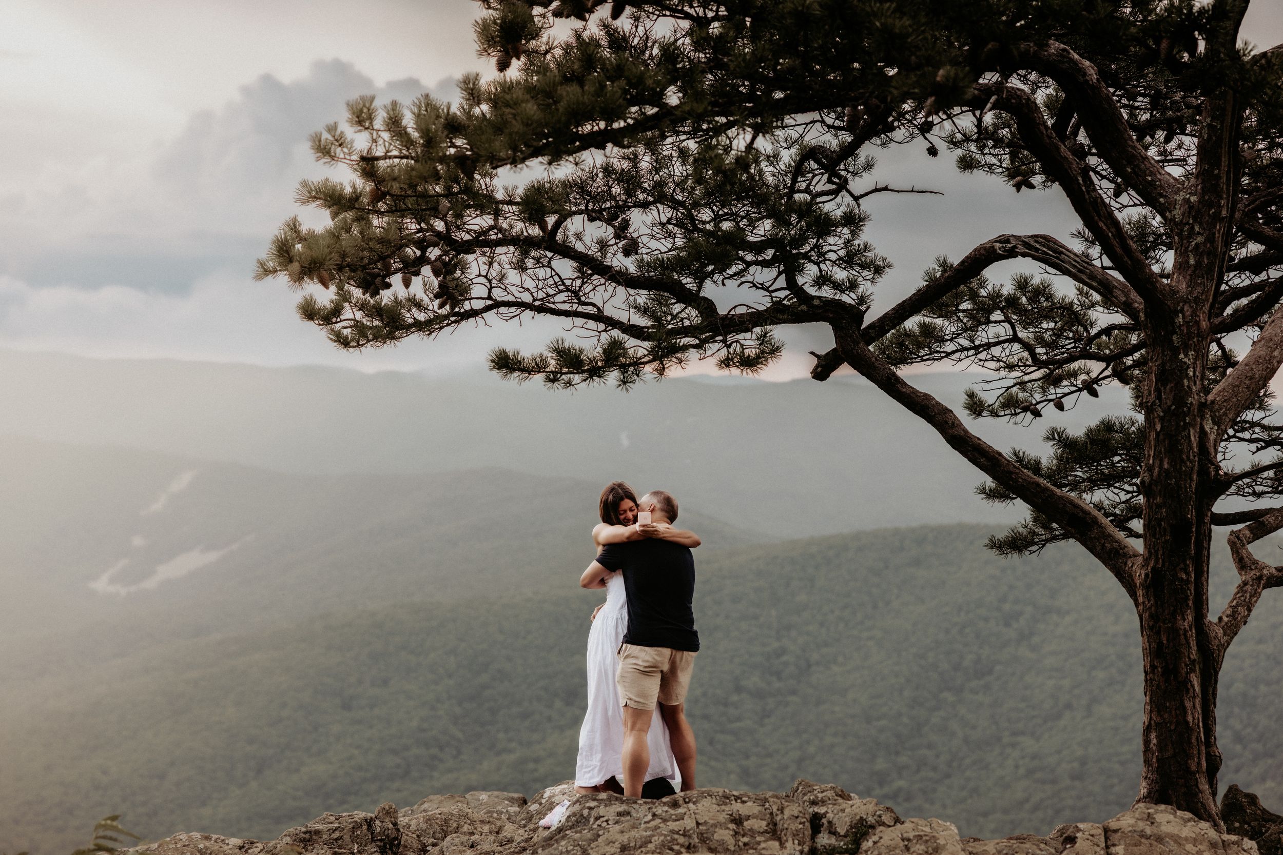 Two silhouettes embrace while standing on a rocky overlook beside a lone pine tree with mountain views in the distance.