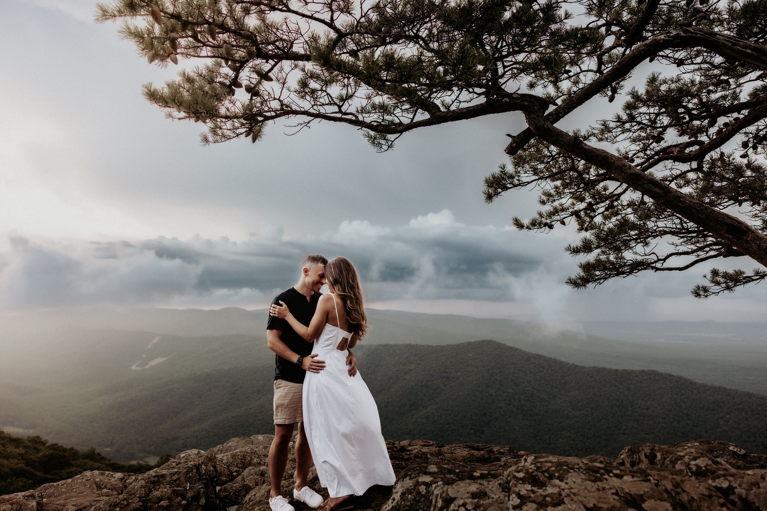 Couple embraces during sunset engagement photo session in foggy mountains with dramatic cloudy sky backdrop.