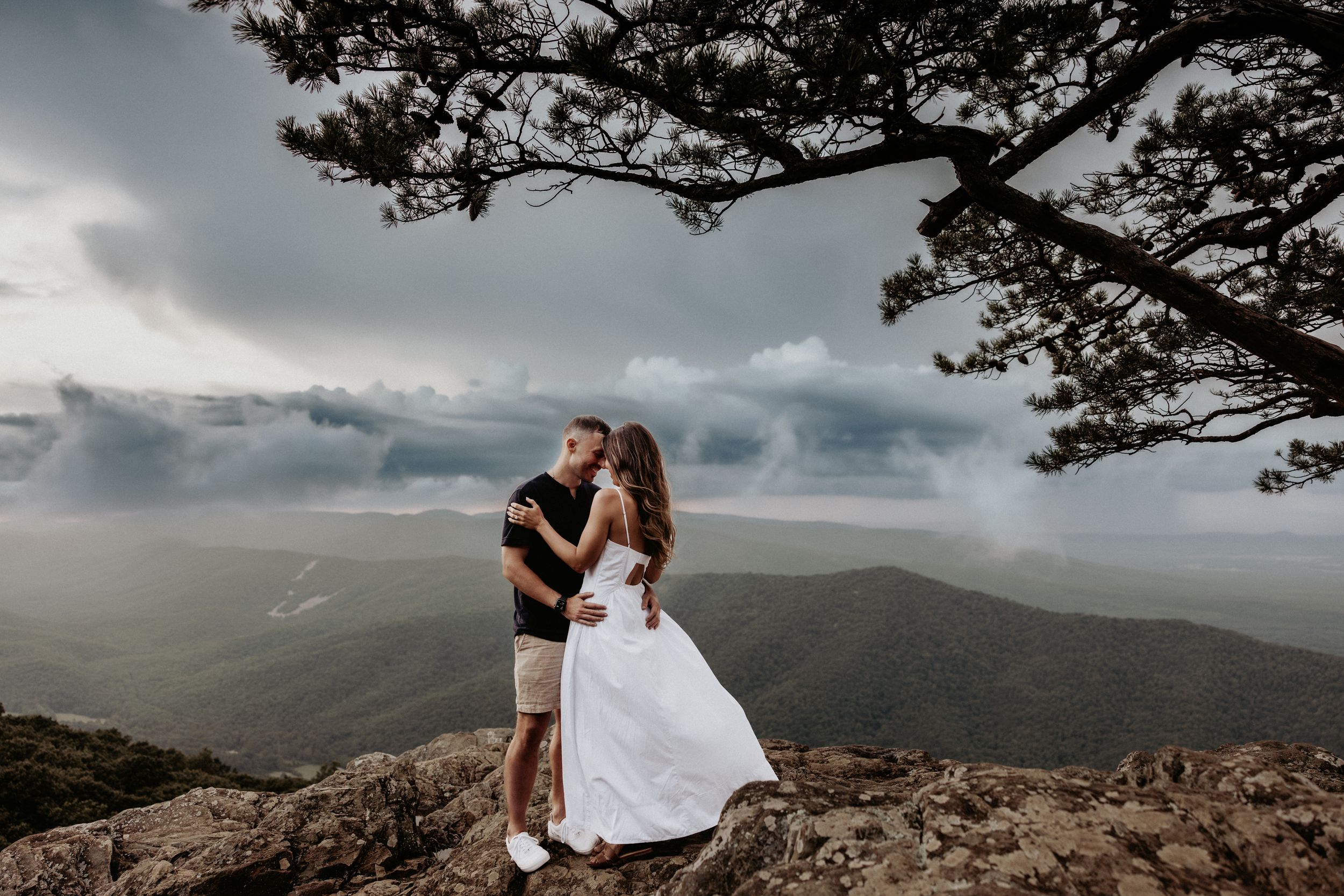 A couple shares an intimate moment on a mountain cliff edge overlooking a dramatic cloudy valley landscape.