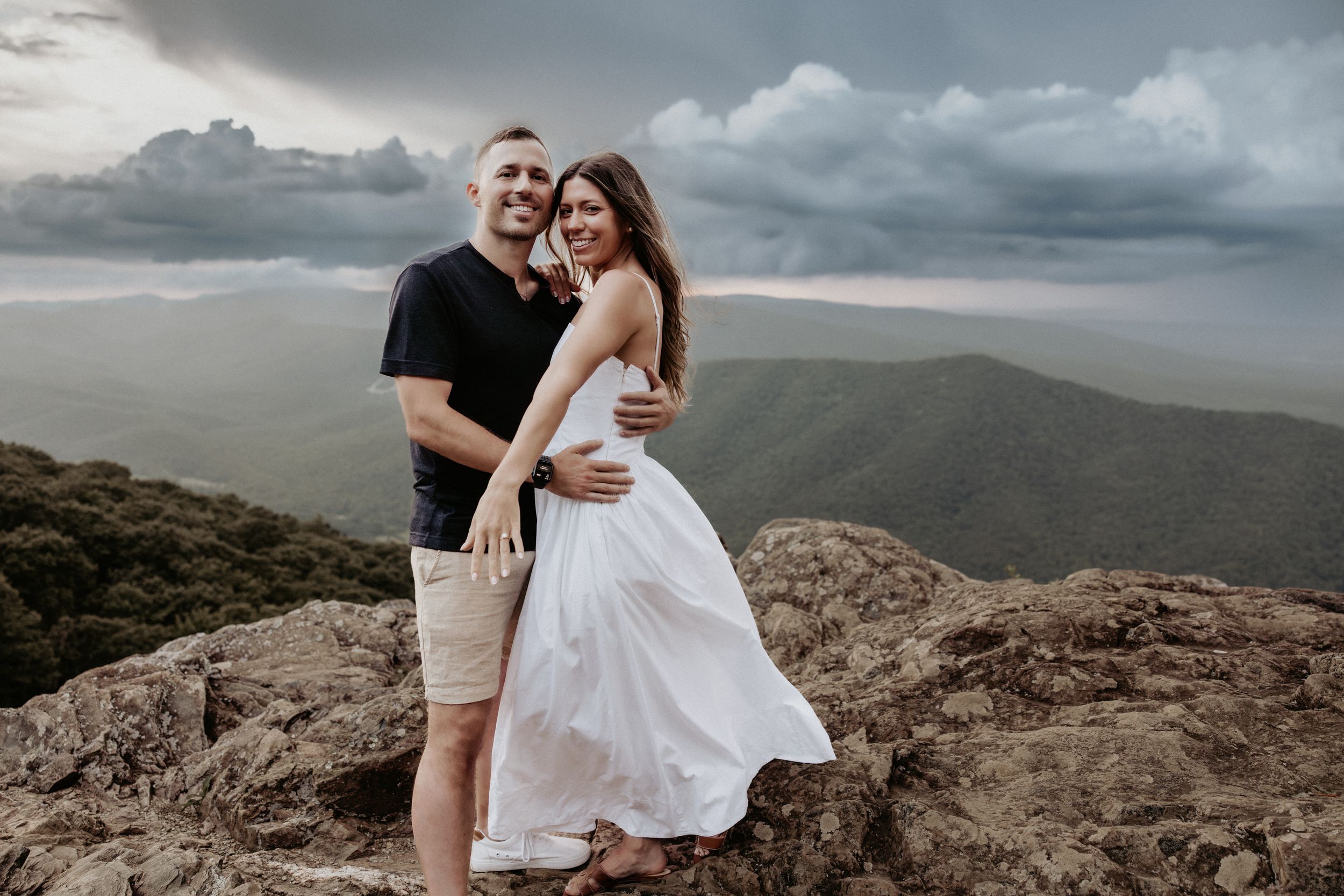 A couple embraces against a dramatic mountain landscape with stormy clouds overhead during a mountaintop photoshoot.