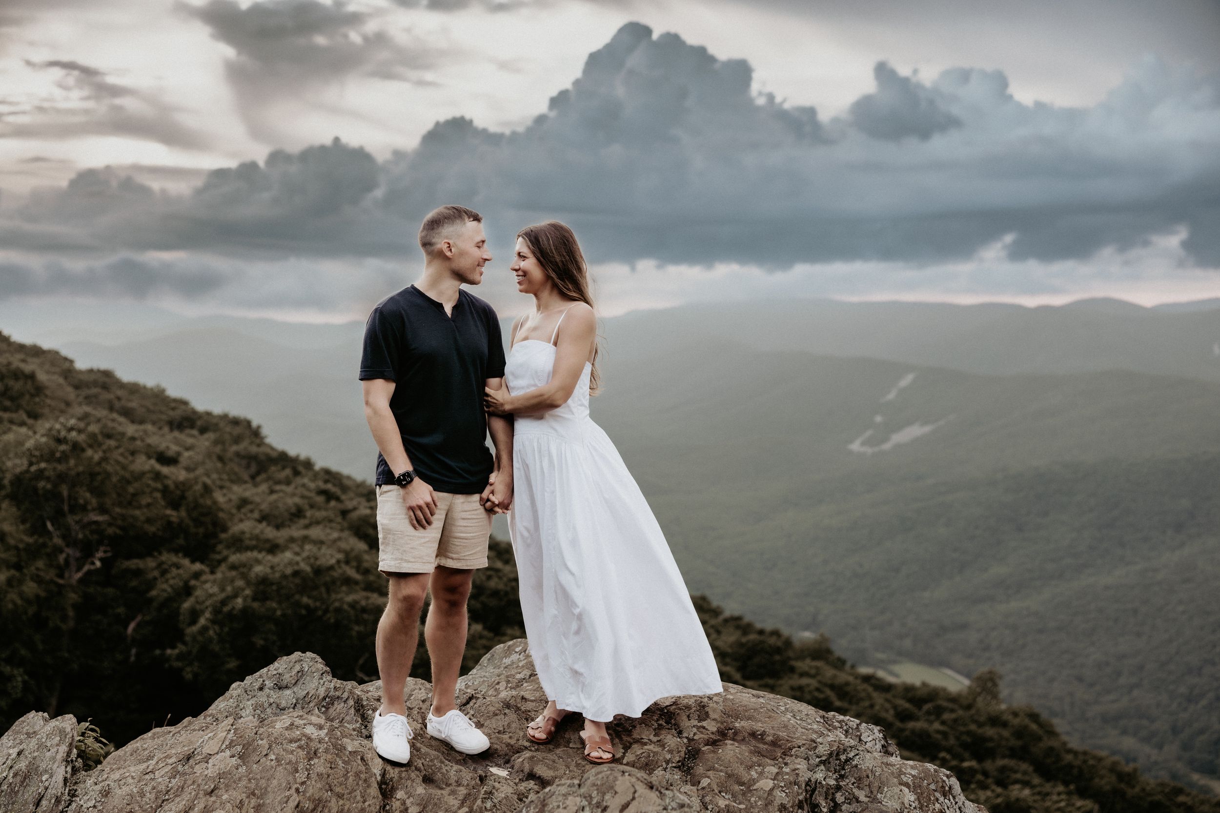 A romantic couple shares an intimate moment atop a mountain peak with dramatic clouds and rolling landscapes in the background.