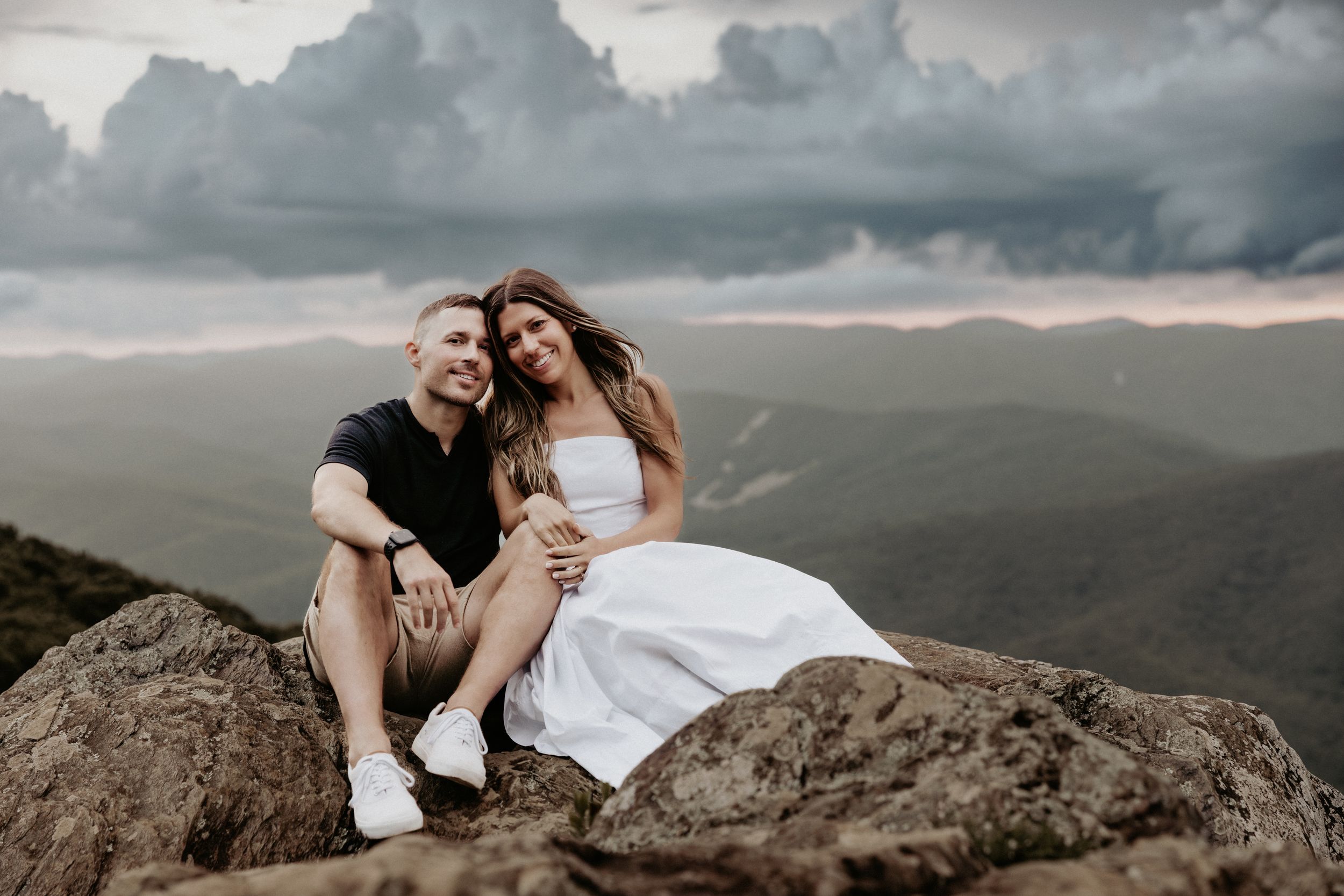 A romantic couple sits intimately on a mountaintop rock overlooking misty mountains during sunset under dramatic clouds.