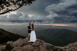 Two figures stand atop a rocky mountain overlook during a dramatic stormy sunset with rolling clouds in the distance.