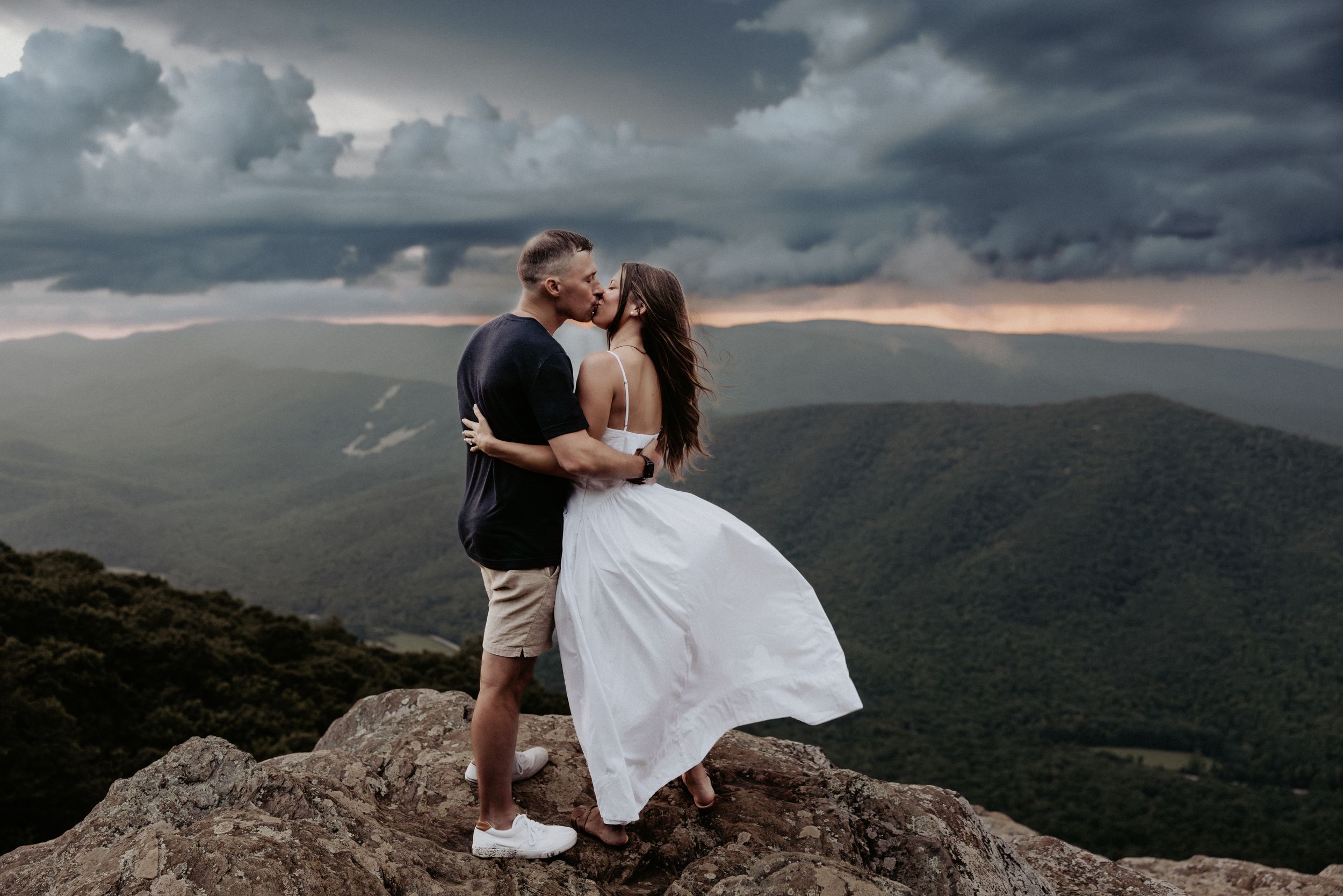 A romantic couple shares a kiss on a mountain summit against dramatic stormy clouds and rolling hills at sunset.