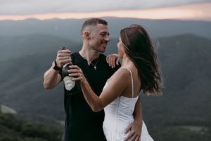 A romantic couple shares drinks while embracing on a mountaintop during sunset against a misty mountain backdrop.