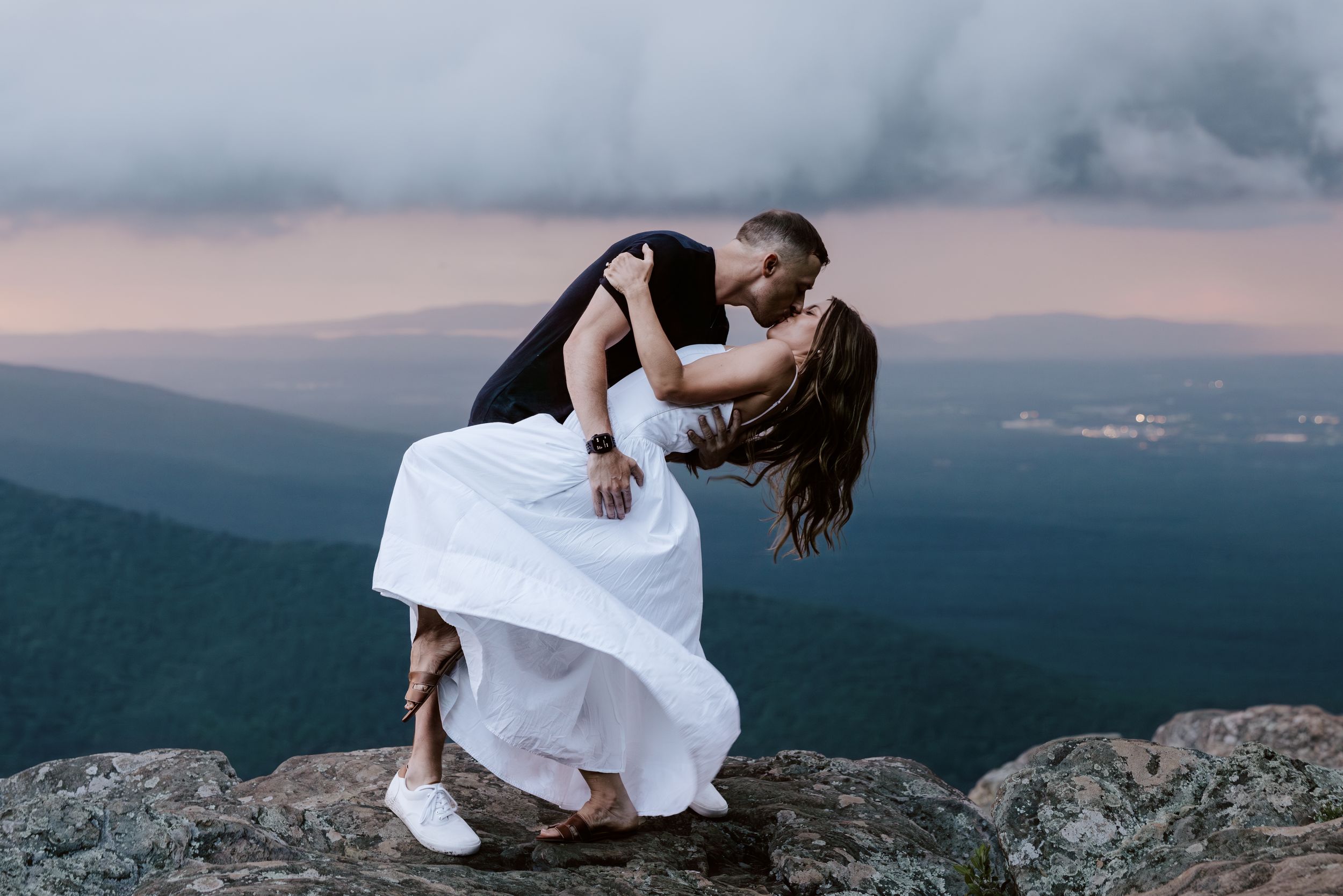 Wedding photos taken on top of a mountain during sunset show embraces and flowing white dresses against dramatic landscapes.