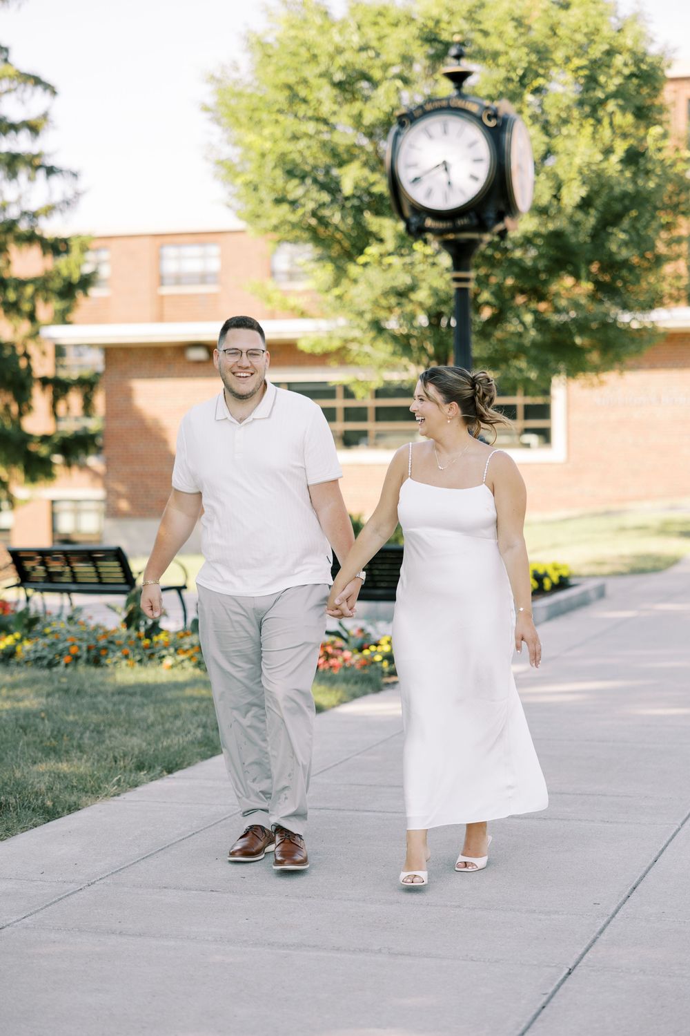 Couple walking together on a campus sidewalk by a decorative clock tower and brick building on a sunny day.