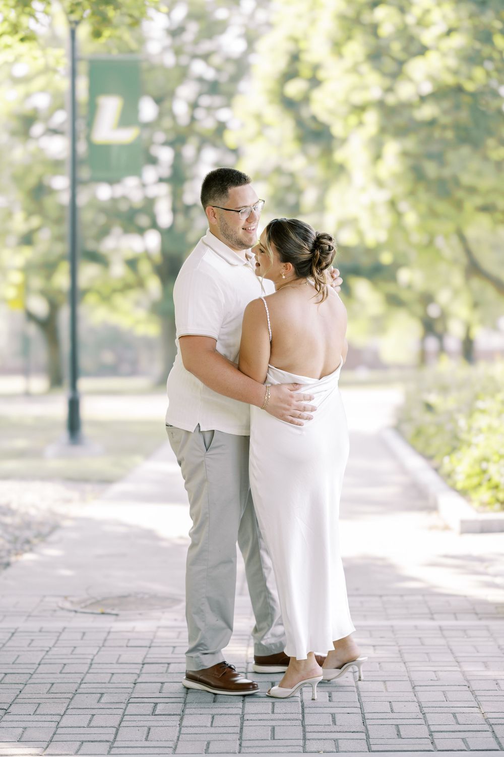 A romantic couple in wedding attire embraces while standing on brick pavement surrounded by greenery in a park setting.