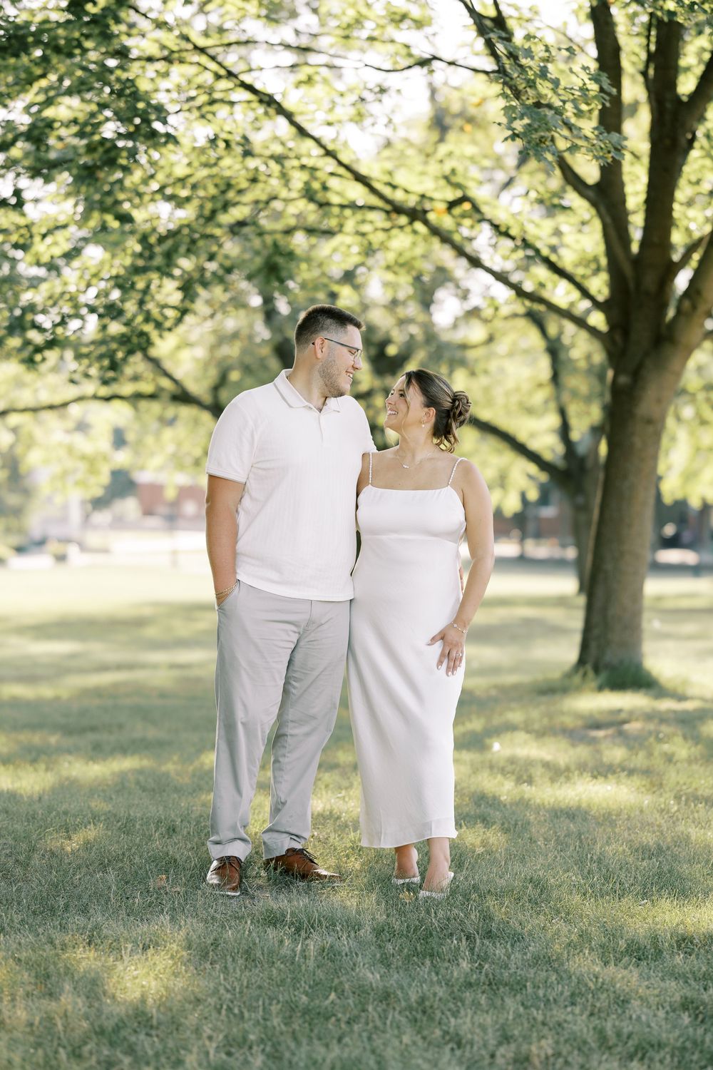A couple in white outfits embraces while standing in a sunny park with tall trees in the background.