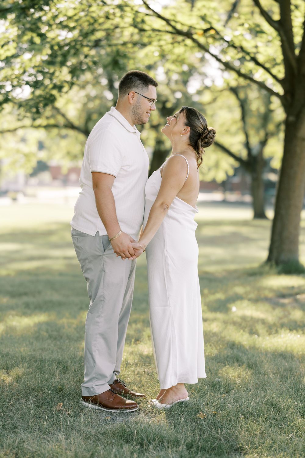 Couple dressed in white clothing share a romantic moment on a sunny day in a park with lush green trees.
