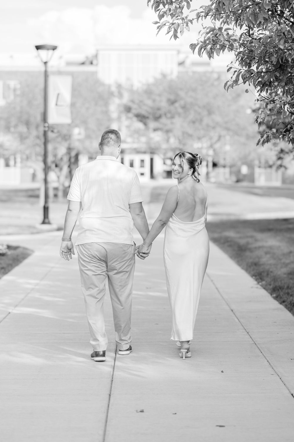 A couple in elegant white attire holding hands while walking down a sidewalk in a black and white photograph.
