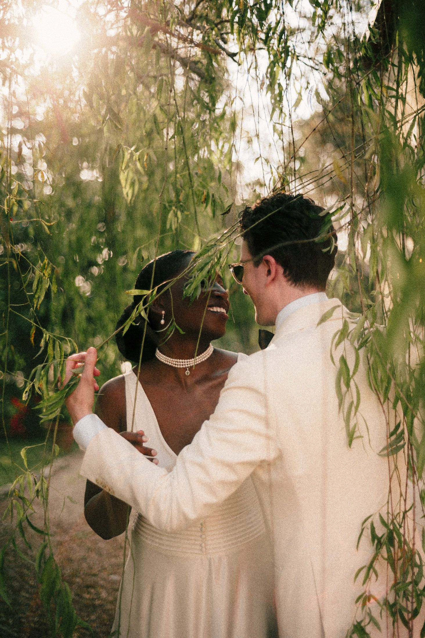 A romantic wedding moment under hanging willow branches with soft sunlight filtering through the leaves.