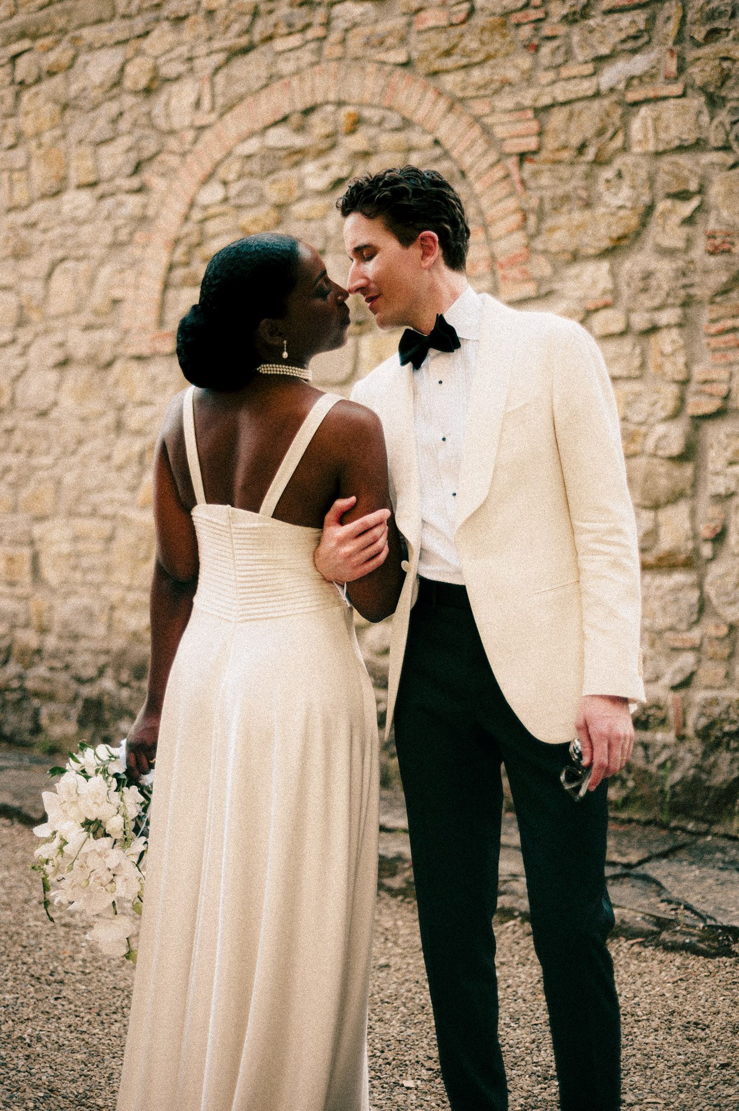 A couple in formal wedding attire shares an intimate moment against a rustic stone wall background.