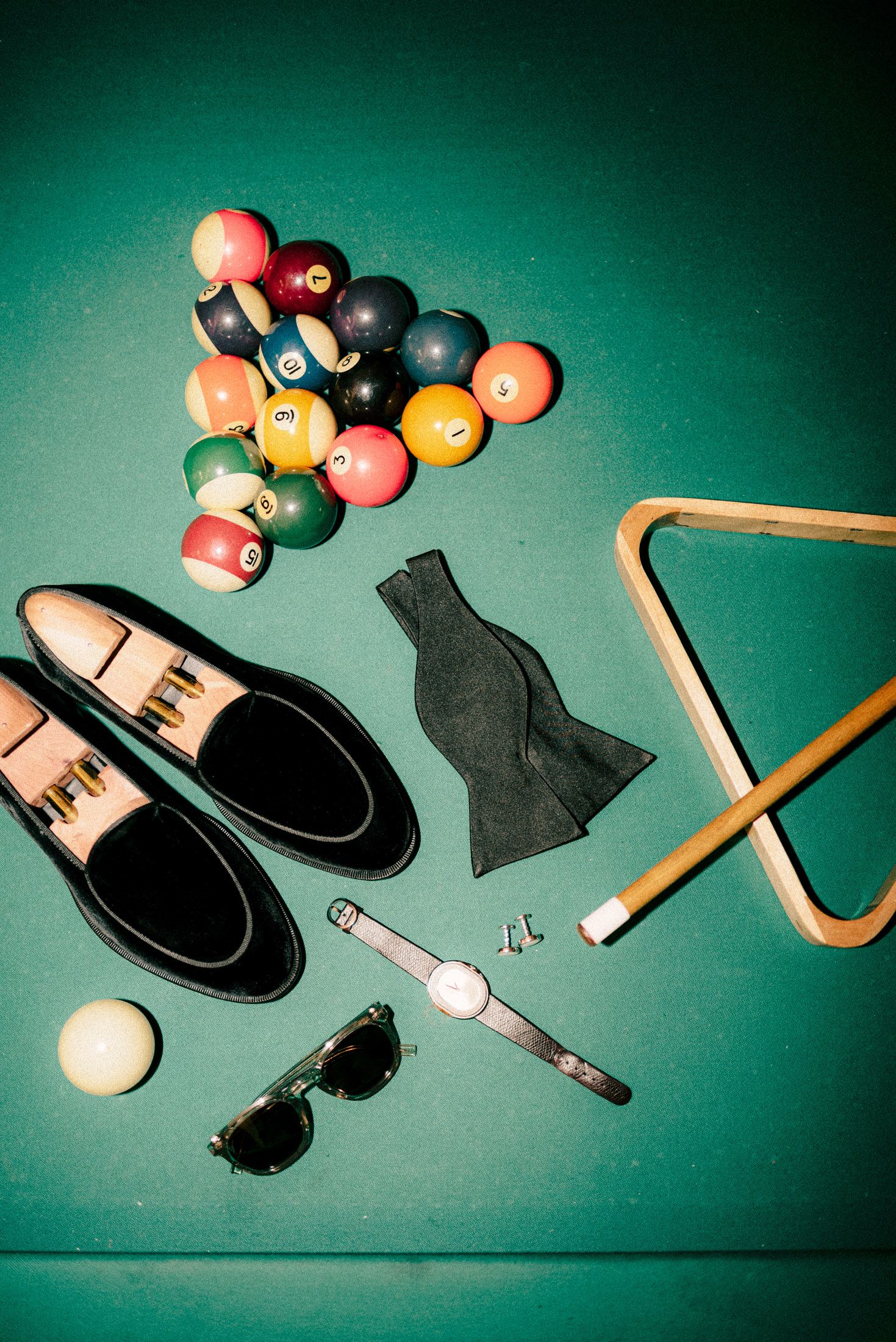 Still life composition of scattered pool balls, triangle rack, chalk and other billiard accessories on green felt background.
