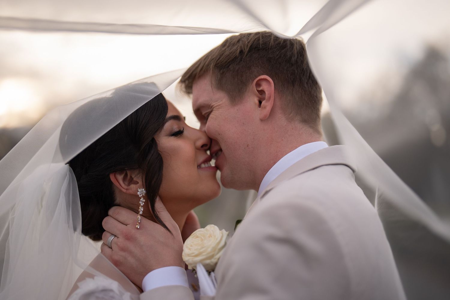 Bride and groom share a romantic kiss under a flowing veil during their outdoor wedding ceremony.