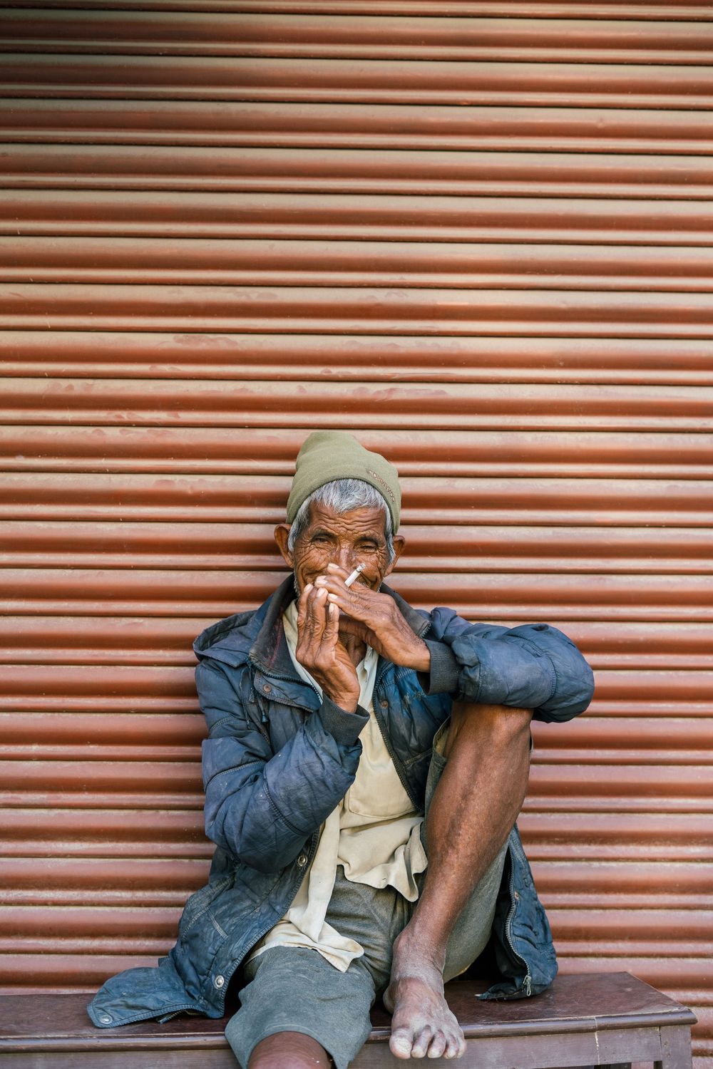 Person sitting against orange shutters wearing denim jacket and beanie in candid street scene.